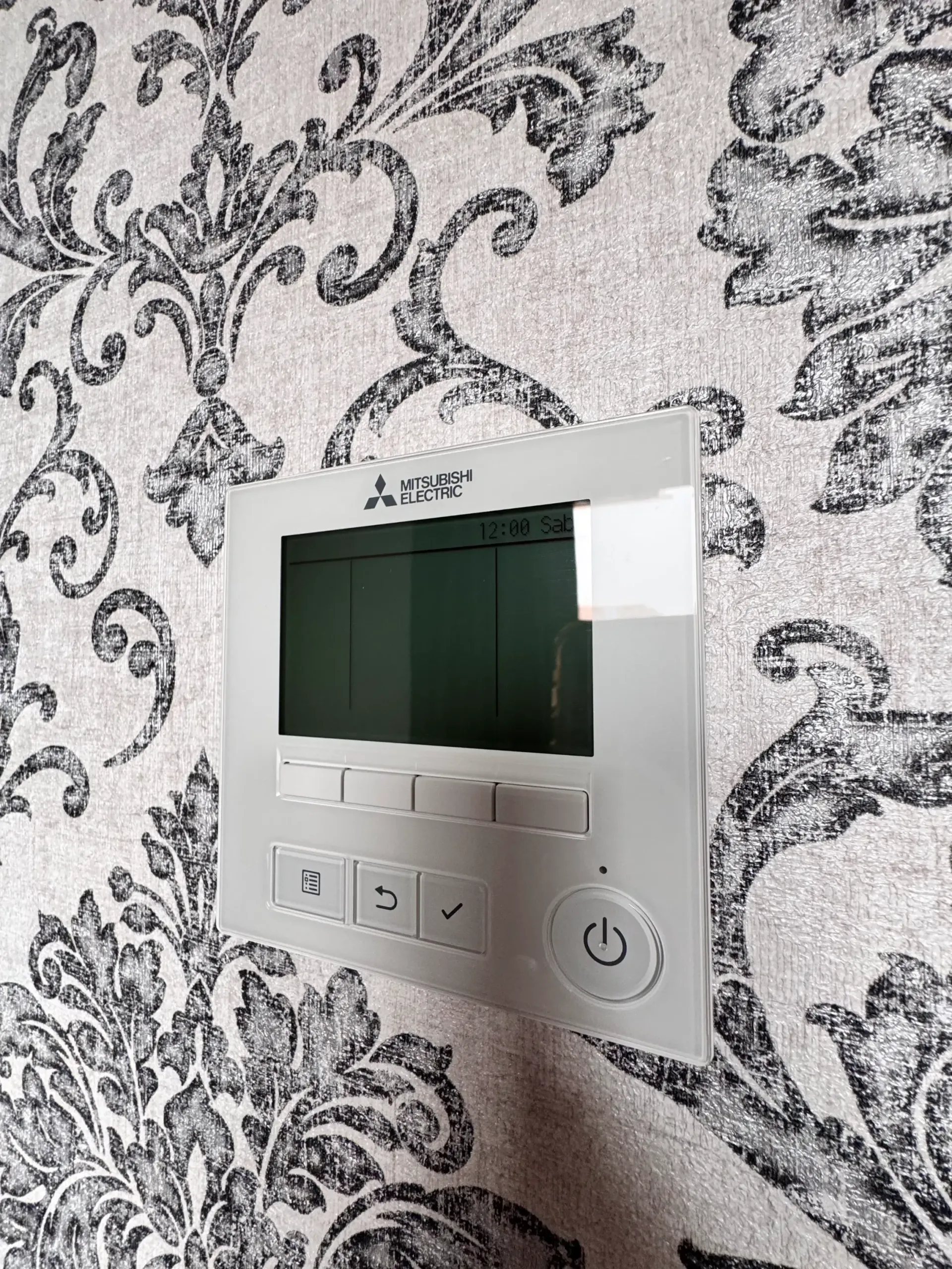 Un controlador de aire acondicionado Mitsubishi blanco montado en una pared con papel tapiz floral estampado en blanco y negro.