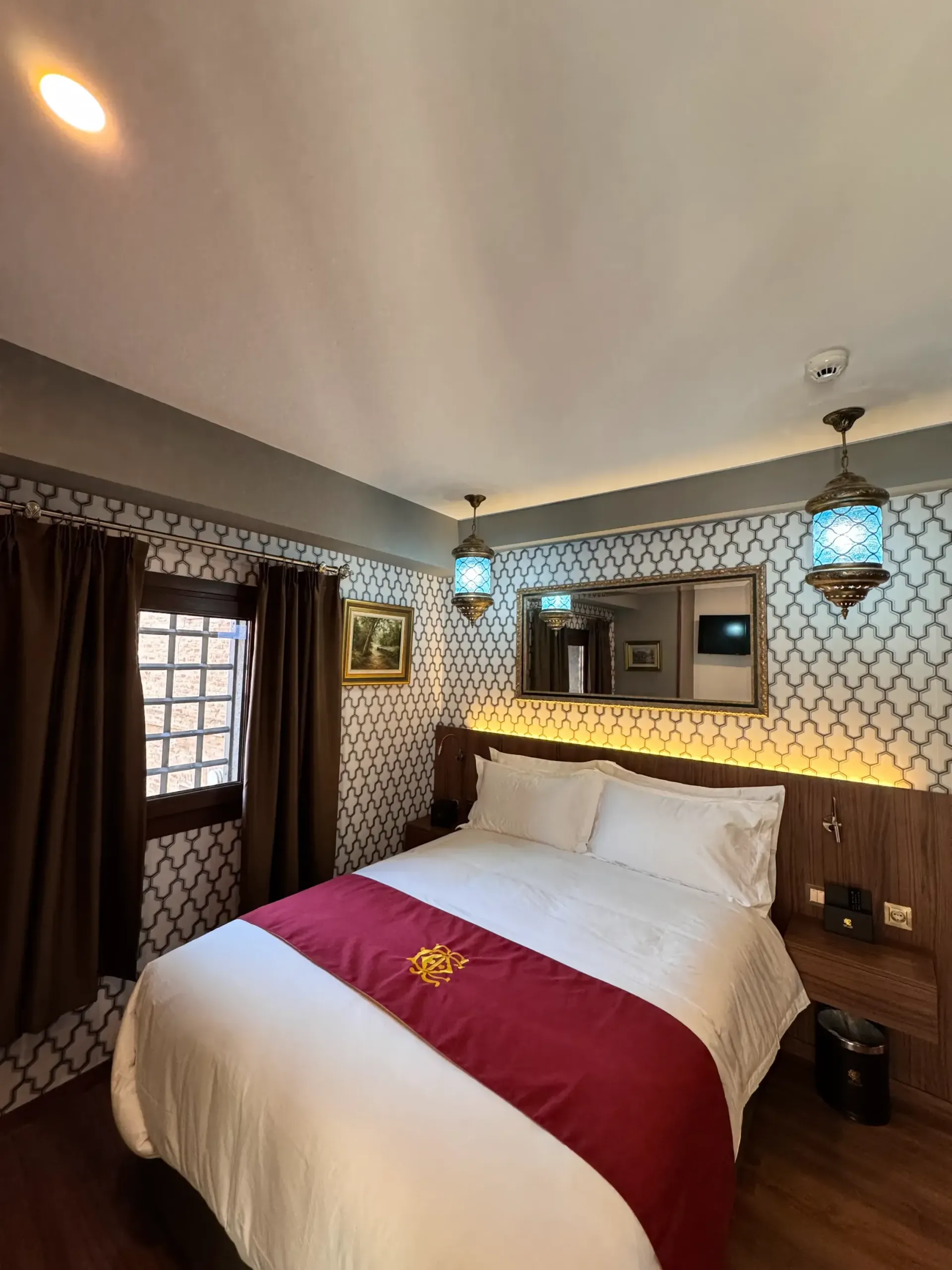 Un dormitorio acogedor con una cama de matrimonio con sábanas blancas, una alfombra color granate, papel pintado decorativo con estampados y lámparas colgantes.