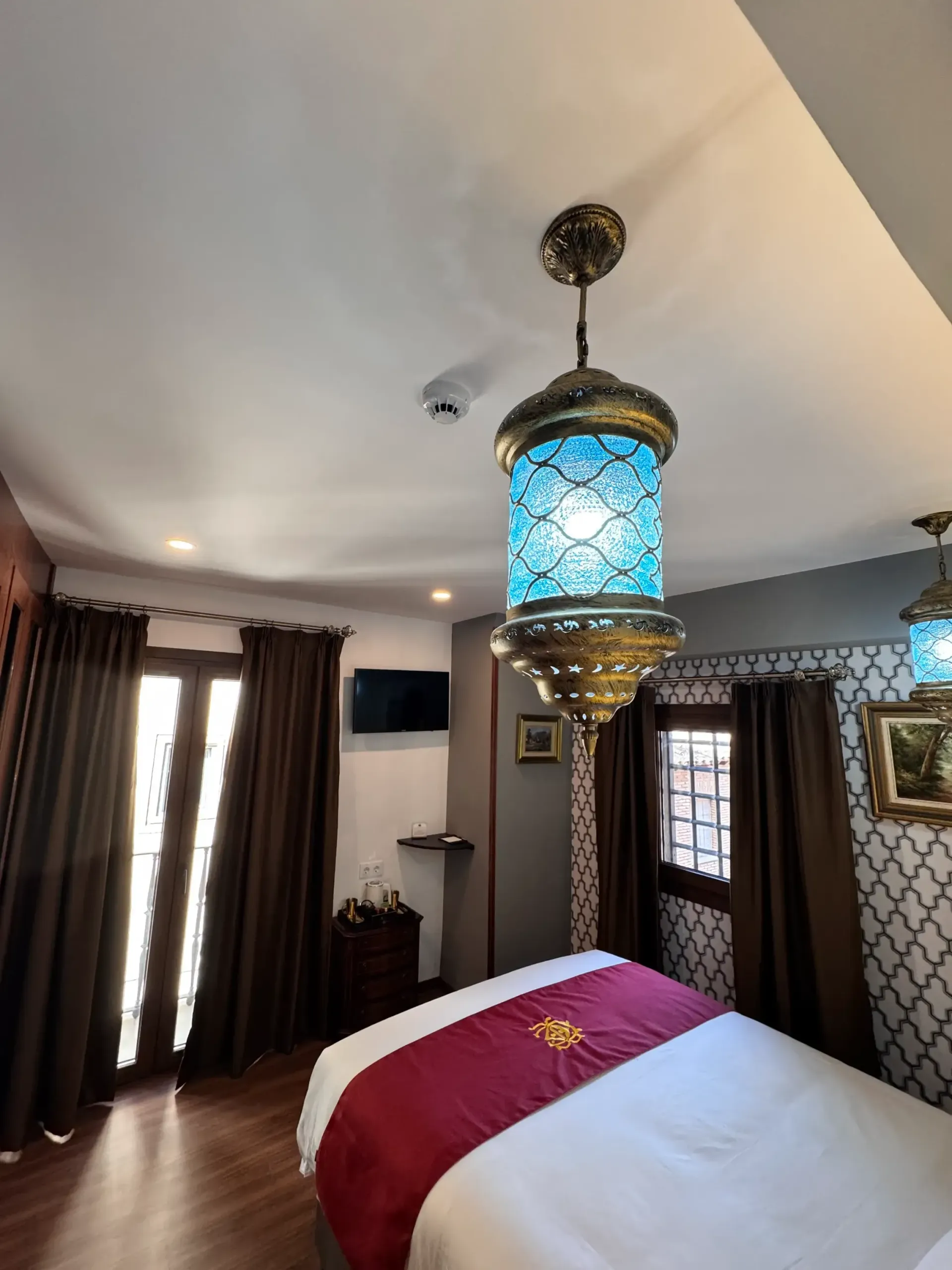 Un dormitorio acogedor con cortinas de color marrón oscuro, una cama con una manta roja y una lámpara colgante azul brillante y ornamentada.