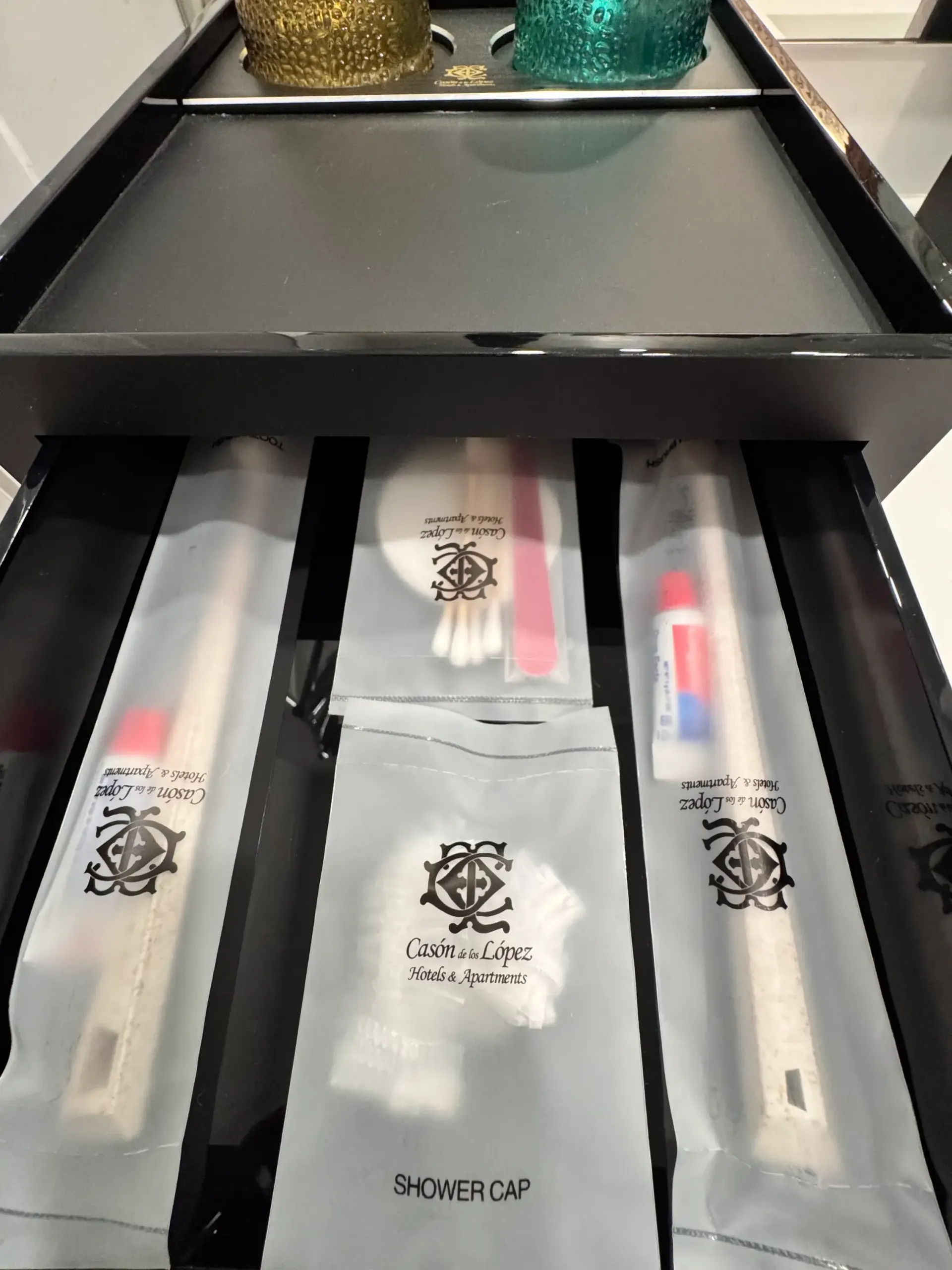 Un cajón abierto que contiene artículos de aseo personal del hotel, envueltos individualmente, entre los que se incluyen cepillos de dientes, bastoncillos de algodón y un gorro de ducha.
