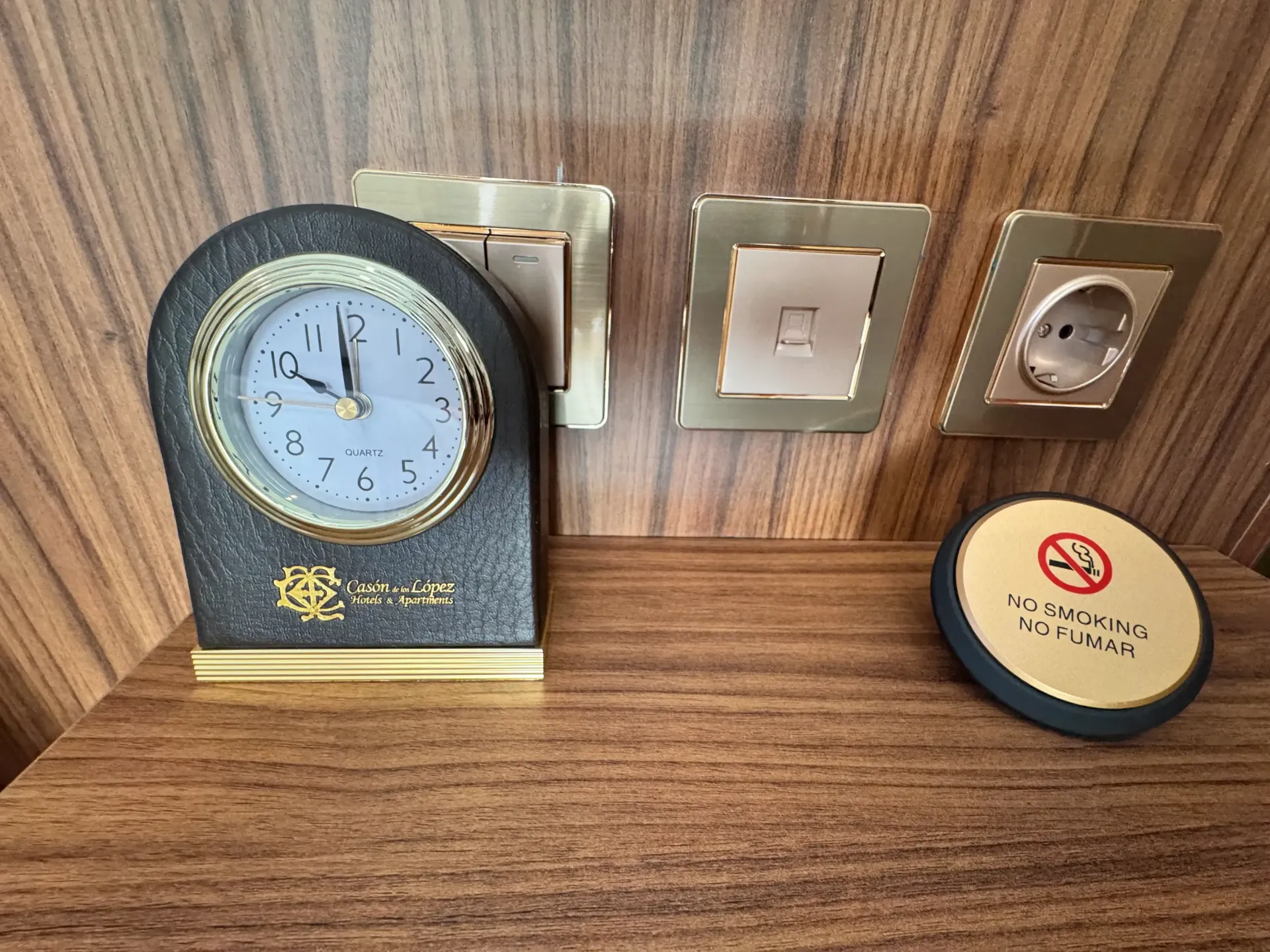 Un reloj analógico de esfera oscura, dos interruptores de luz y una toma de corriente europea sobre un cabecero de madera con un icono de prohibido fumar.