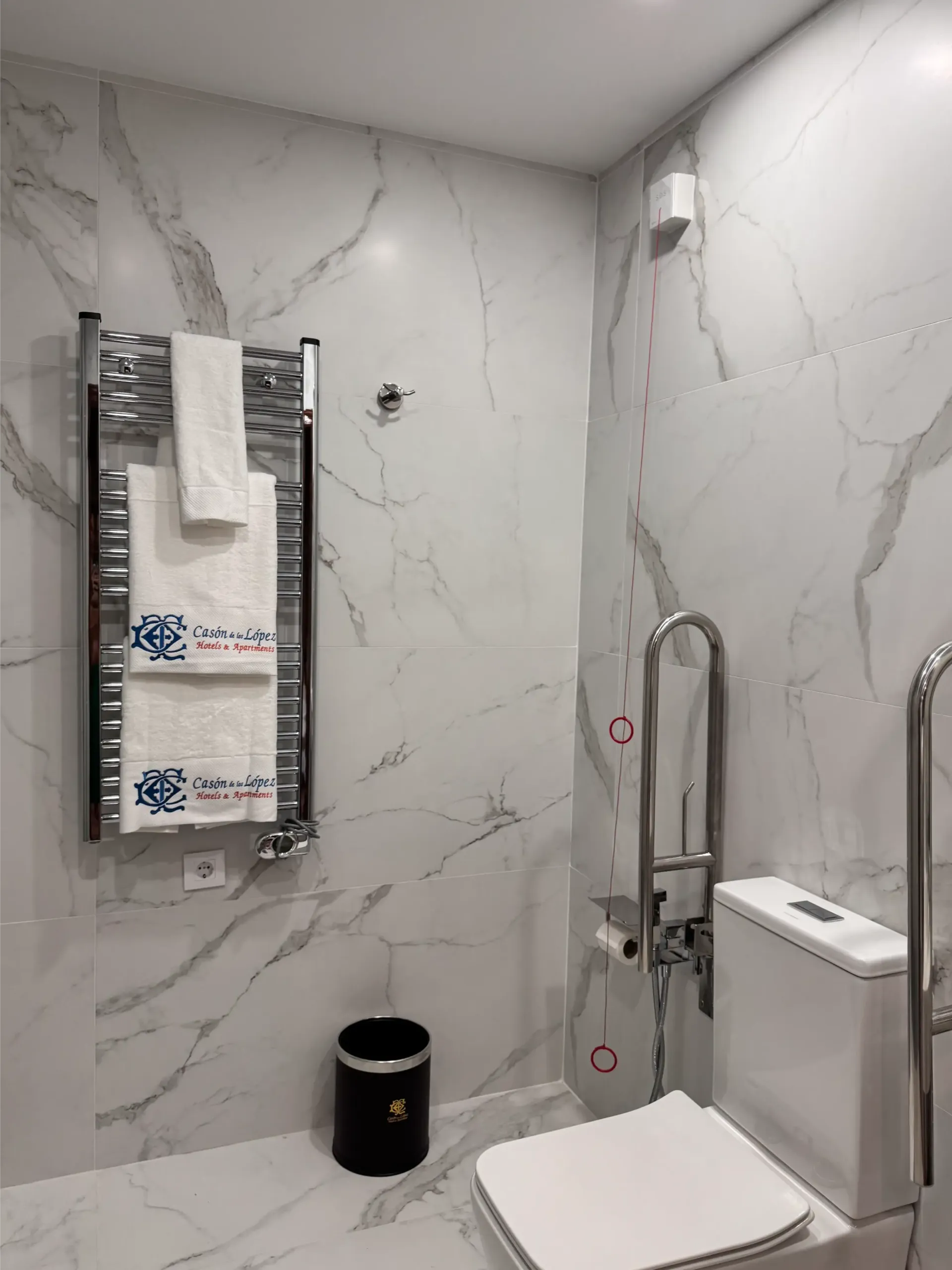 Baño accesible con azulejos que imitan el mármol, toallero eléctrico, papelera negra e inodoro con barras de seguridad.