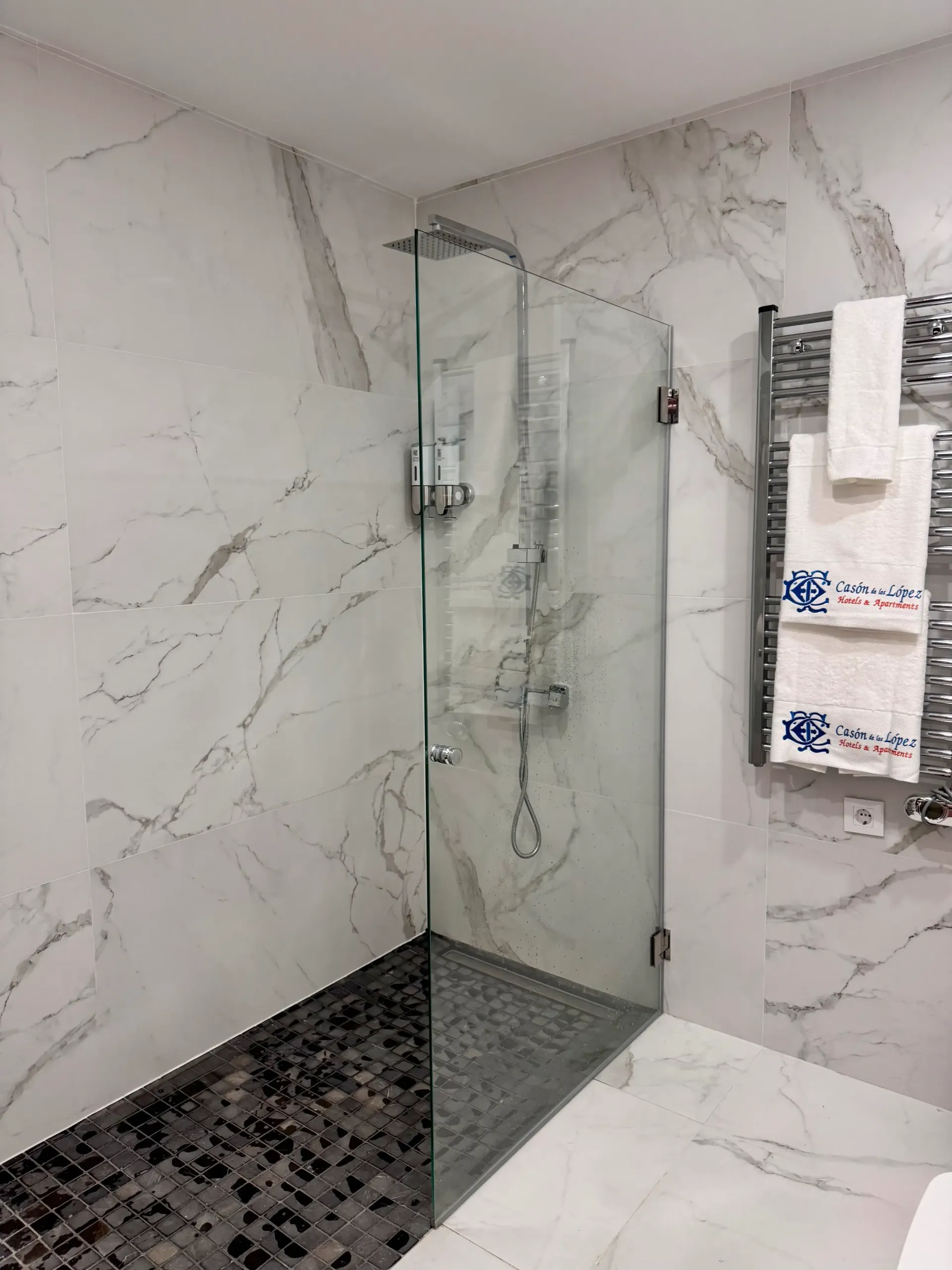 Una ducha con mampara de cristal, suelo de guijarros oscuros, rodeada de azulejos blancos con estampado de mármol y un toallero repleto de toallas.