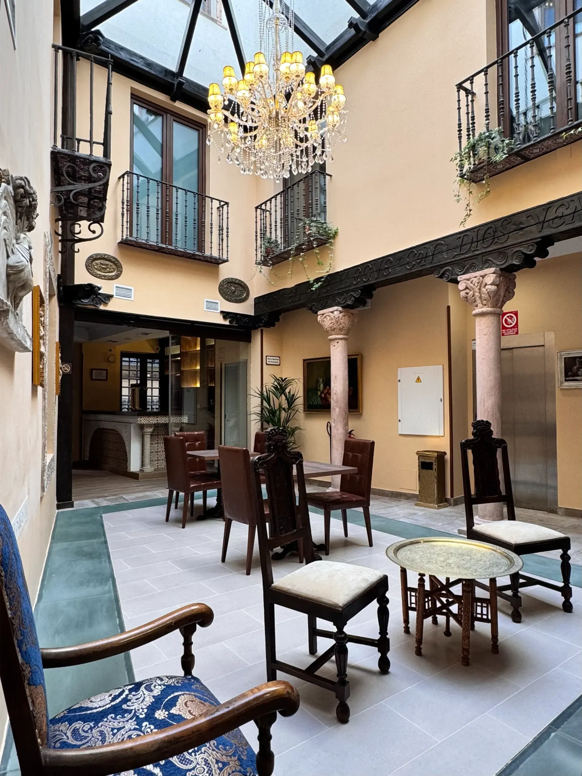 Un patio interior con paredes de color beige, techo de cristal, una gran lámpara de araña y varias sillas de madera alrededor de mesas pequeñas.