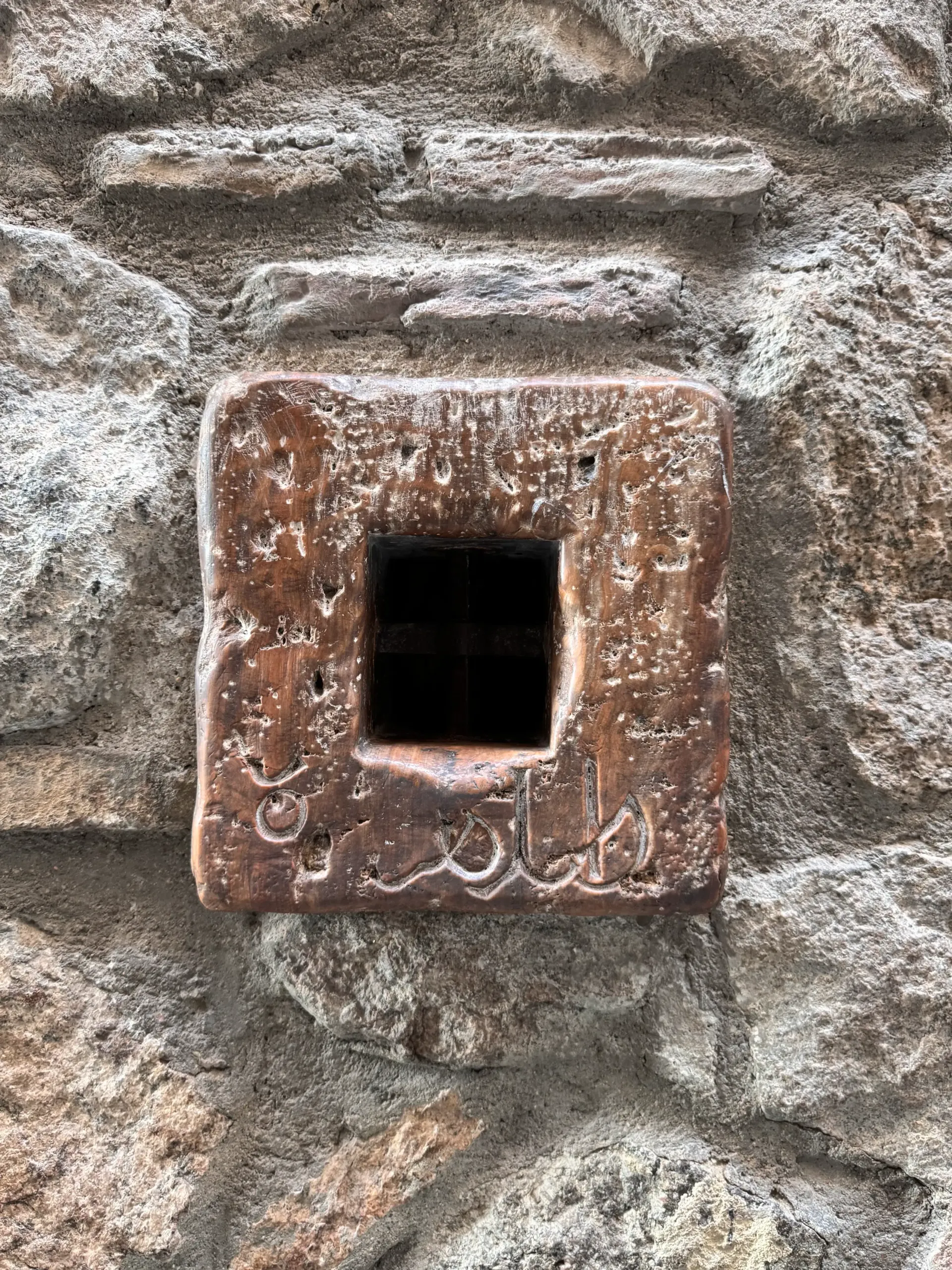Una piedra cuadrada de color marrón con inscripciones árabes talladas que rodea un pequeño agujero rectangular, incrustada en un muro de piedra.