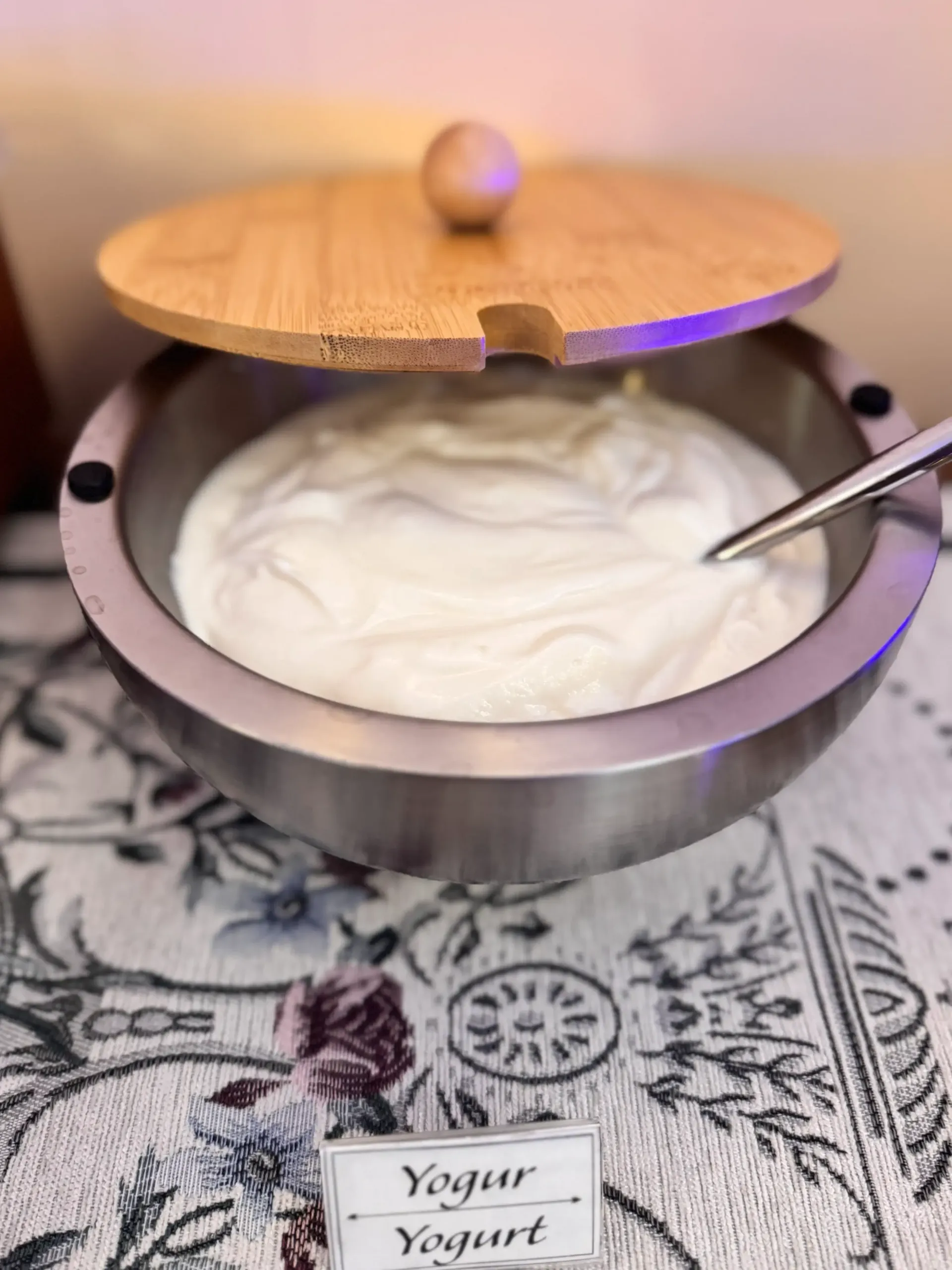Un bol de acero inoxidable con yogur blanco y tapa de madera, sobre un mantel estampado con la etiqueta 
