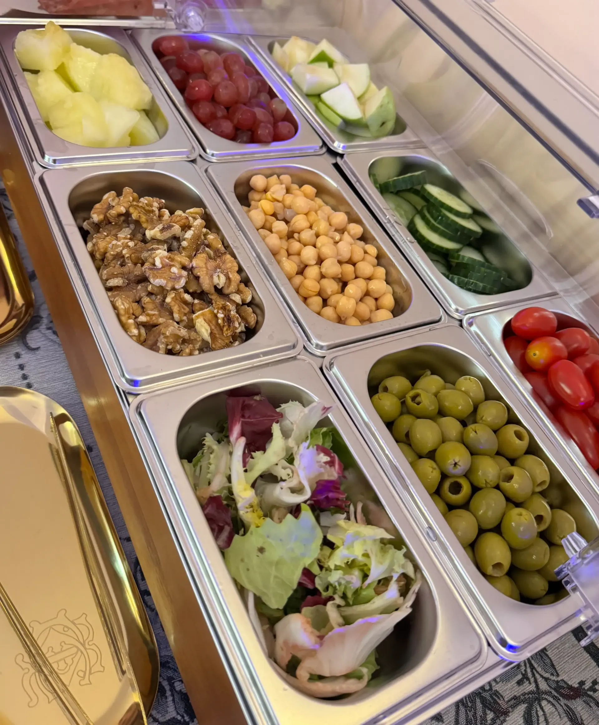 Una barra de ensaladas con recipientes de acero inoxidable que contienen verduras, frutas, garbanzos, nueces y aceitunas variadas.