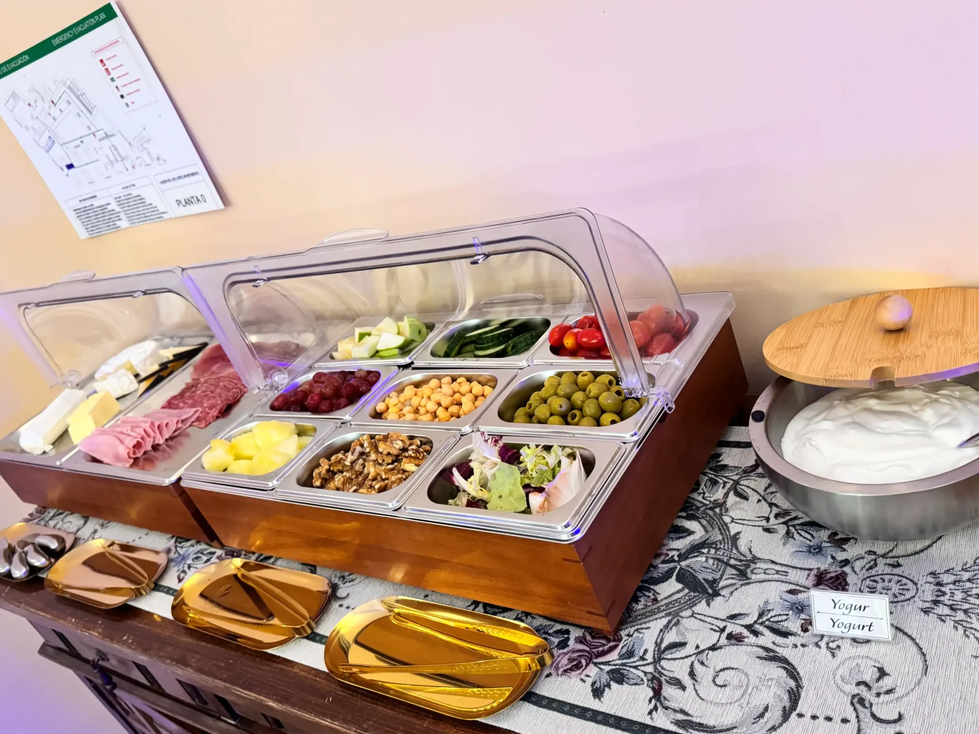 Una barra de ensaladas tipo bufé con varios ingredientes en recipientes metálicos, junto con un cuenco de aderezo, dispuesta sobre un mantel estampado.