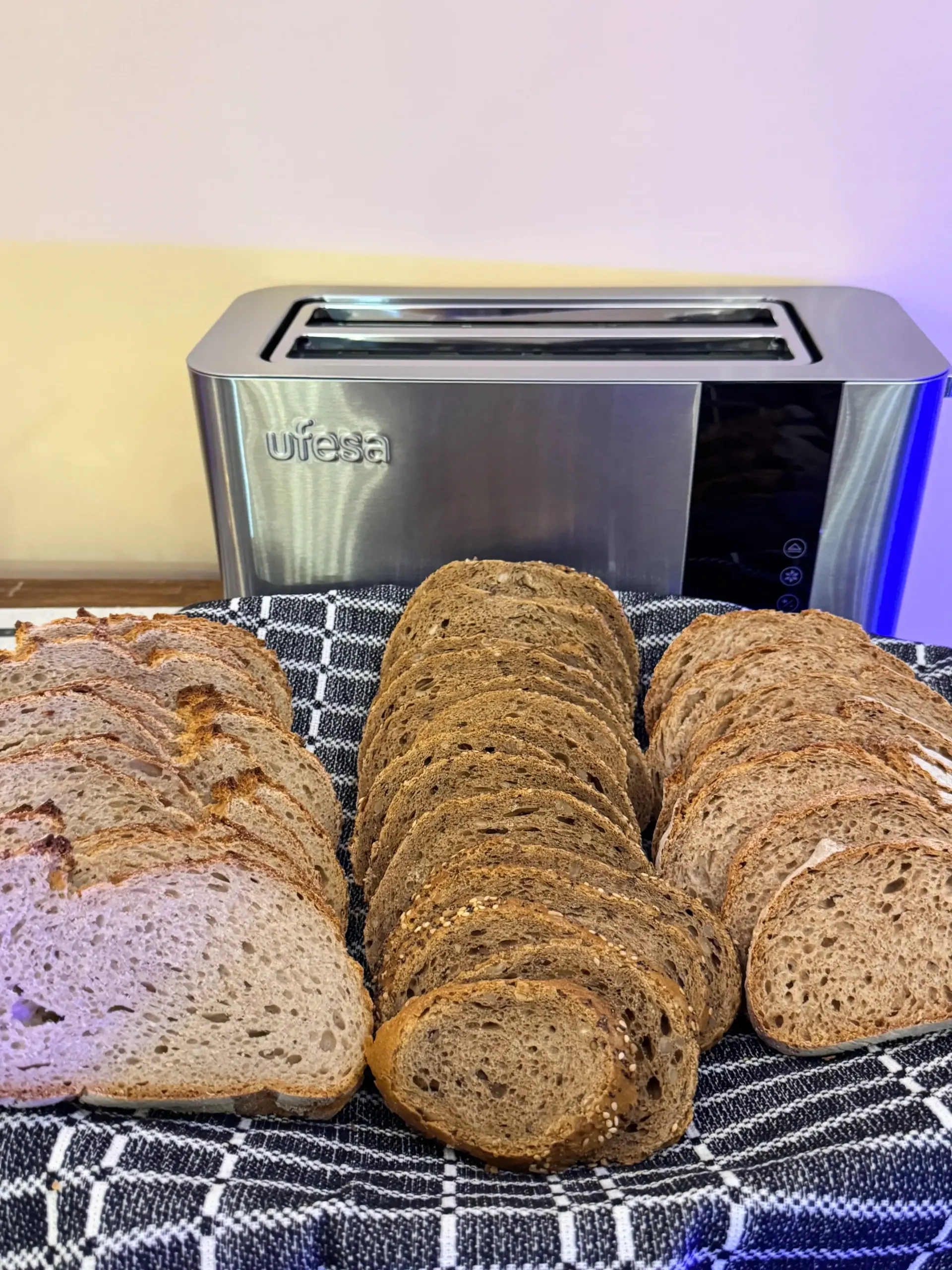 Una tostadora plateada detrás de una cesta con muchas rebanadas de pan integral dispuestas sobre un paño a cuadros blancos y negros.