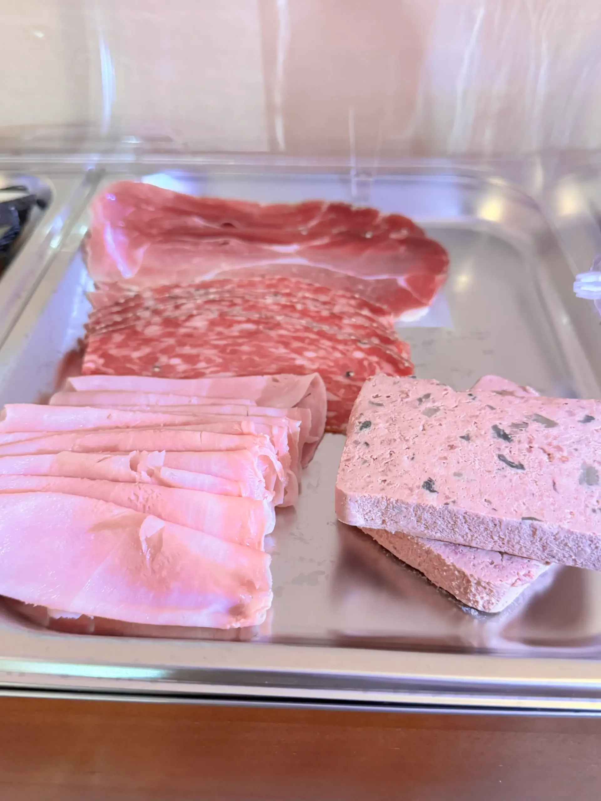 Una bandeja metálica para servir que contiene lonchas de prosciutto, salami, jamón y un paté o terrina de carne.