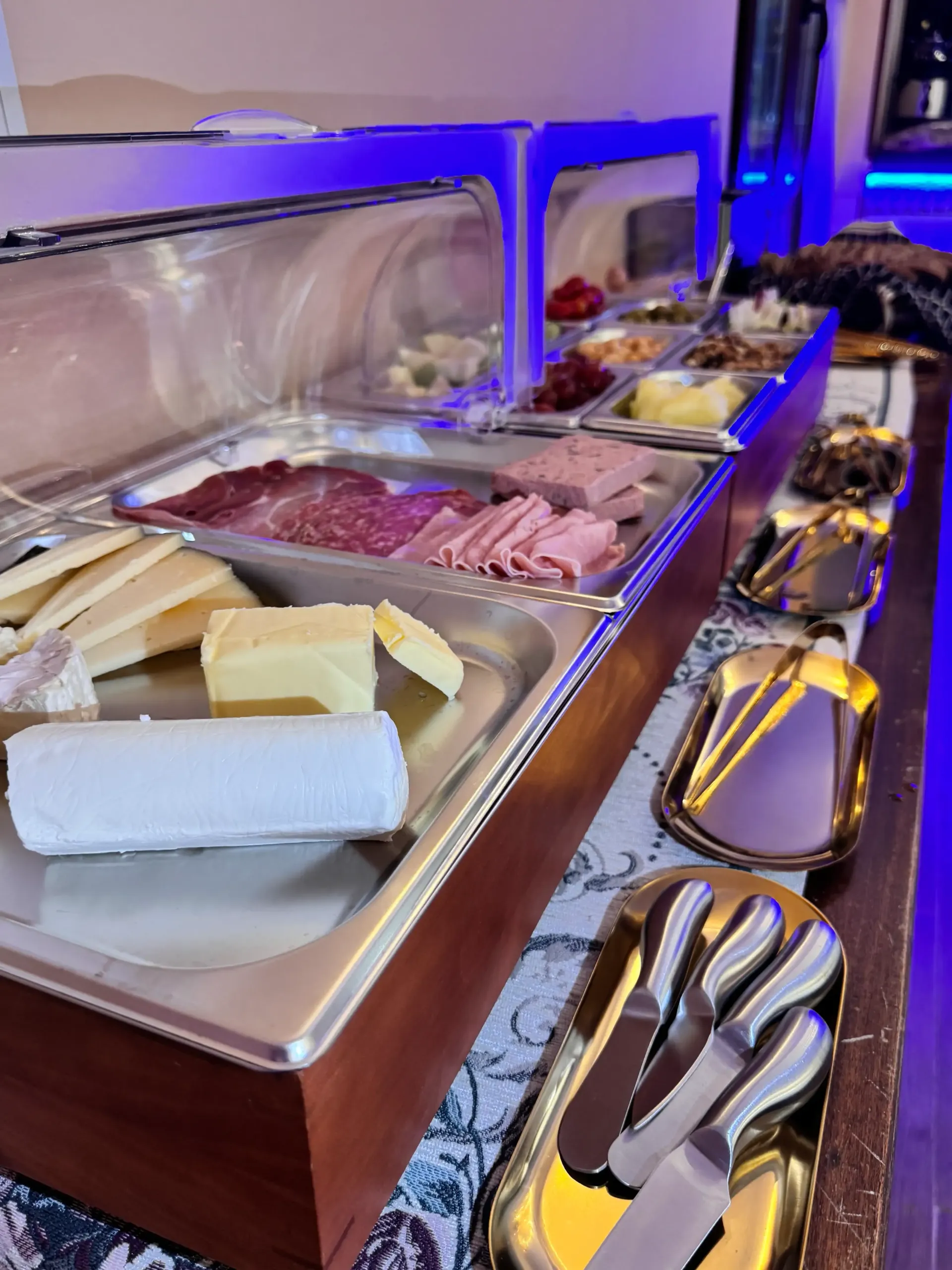 Un bufé con una variedad de quesos, embutidos en lonchas y utensilios para servir, dispuestos sobre una mesa larga.