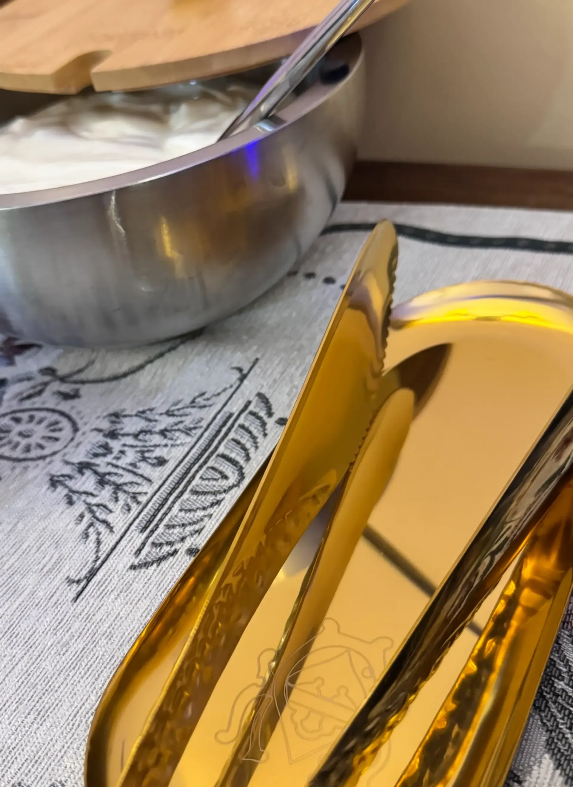 Unas pinzas de servir de color dorado descansan sobre un mantel estampado cerca de un cuenco metálico de yogur.