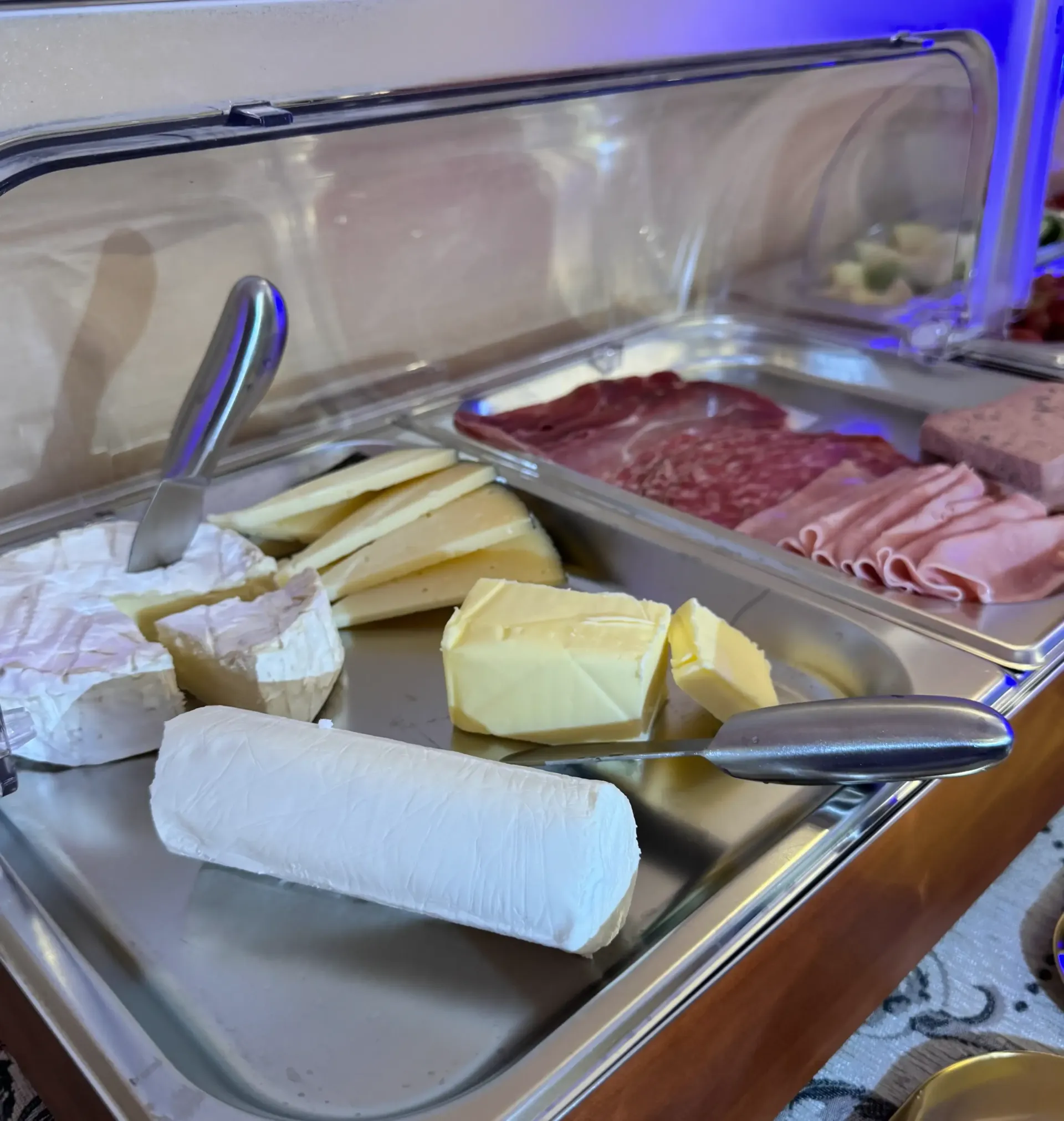 Un bufé con una selección de quesos, embutidos y mantequilla servidos en bandejas de acero inoxidable bajo una vitrina.