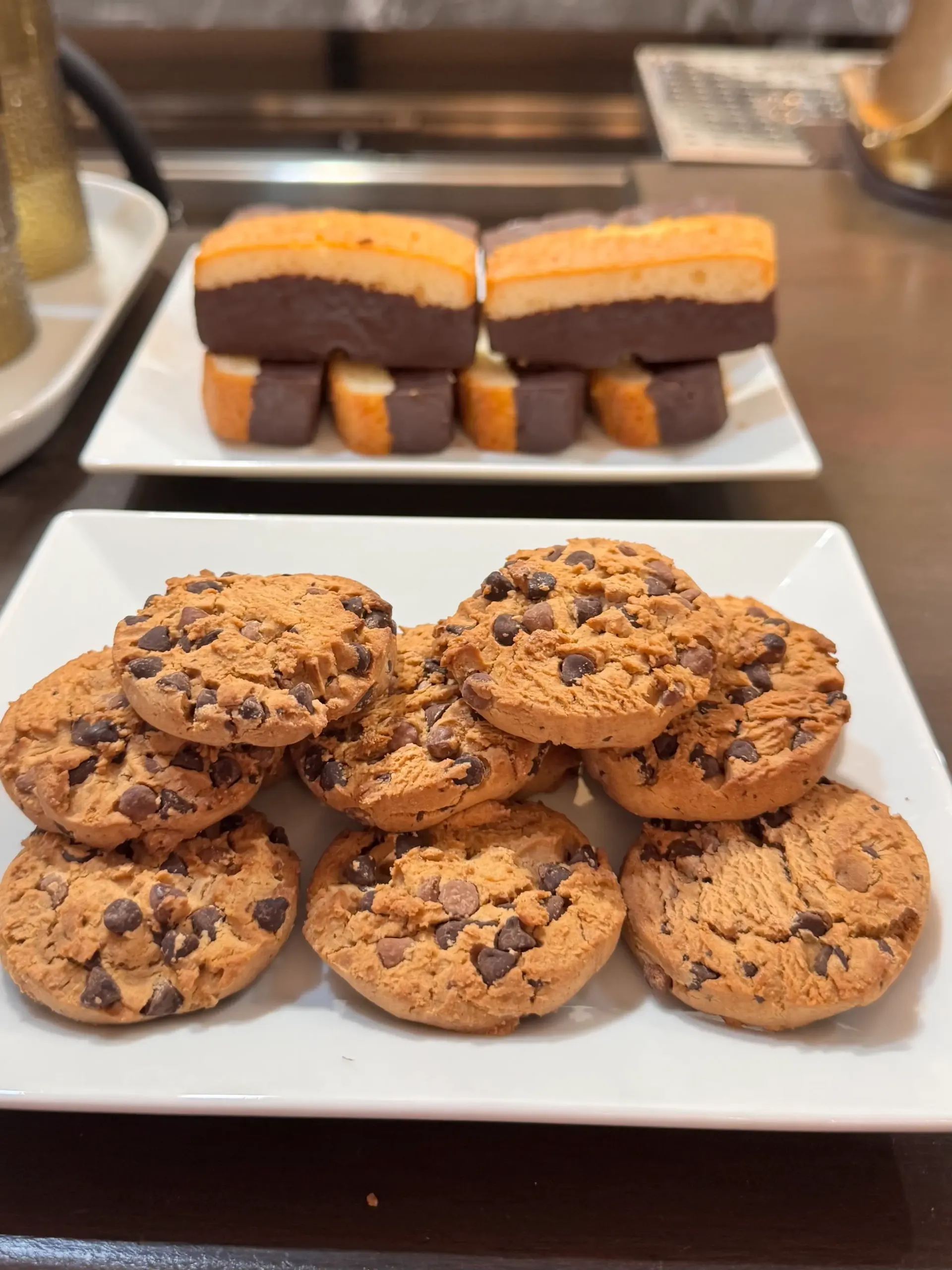 Dos platos blancos sobre una superficie oscura: uno con galletas rectangulares bañadas en chocolate y el otro con galletas con trocitos de chocolate apiladas.