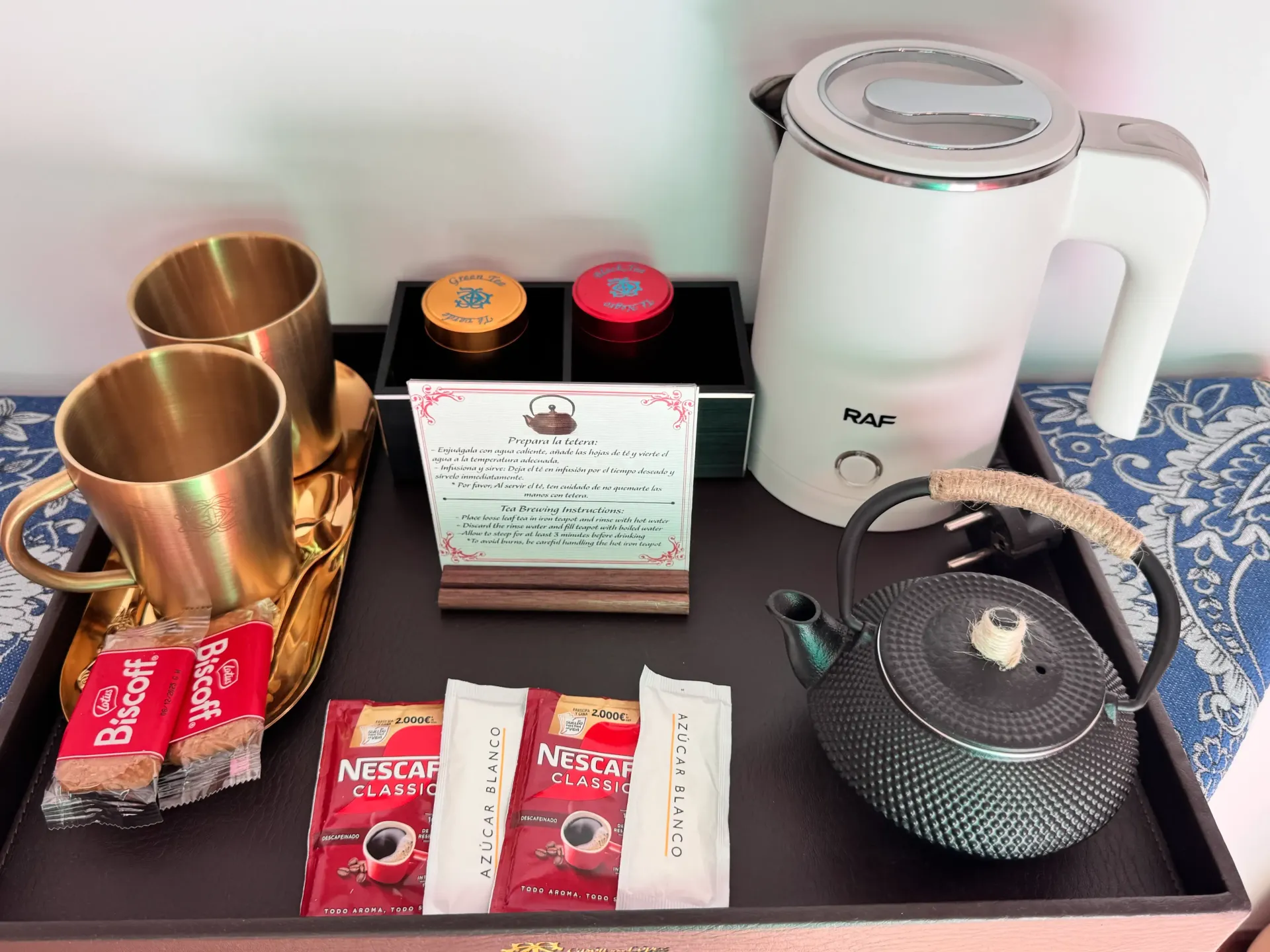 Una bandeja con una tetera eléctrica blanca, una tetera oscura, dos tazas de latón, sobres de café, galletas y un pequeño cartel.