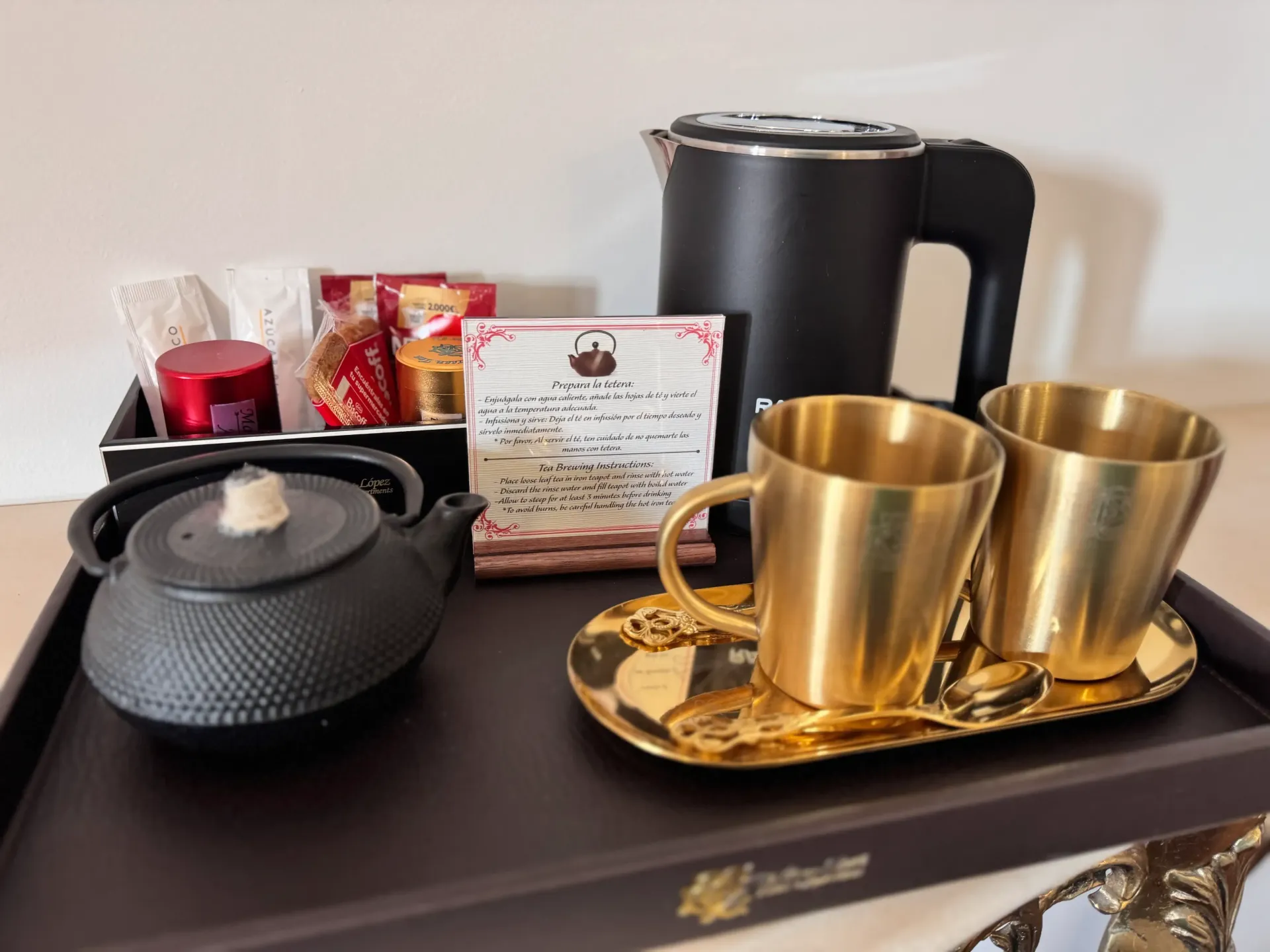 Una bandeja de té de hotel con una tetera negra, una tetera de hierro fundido, dos tazas doradas y paquetes de té surtidos.