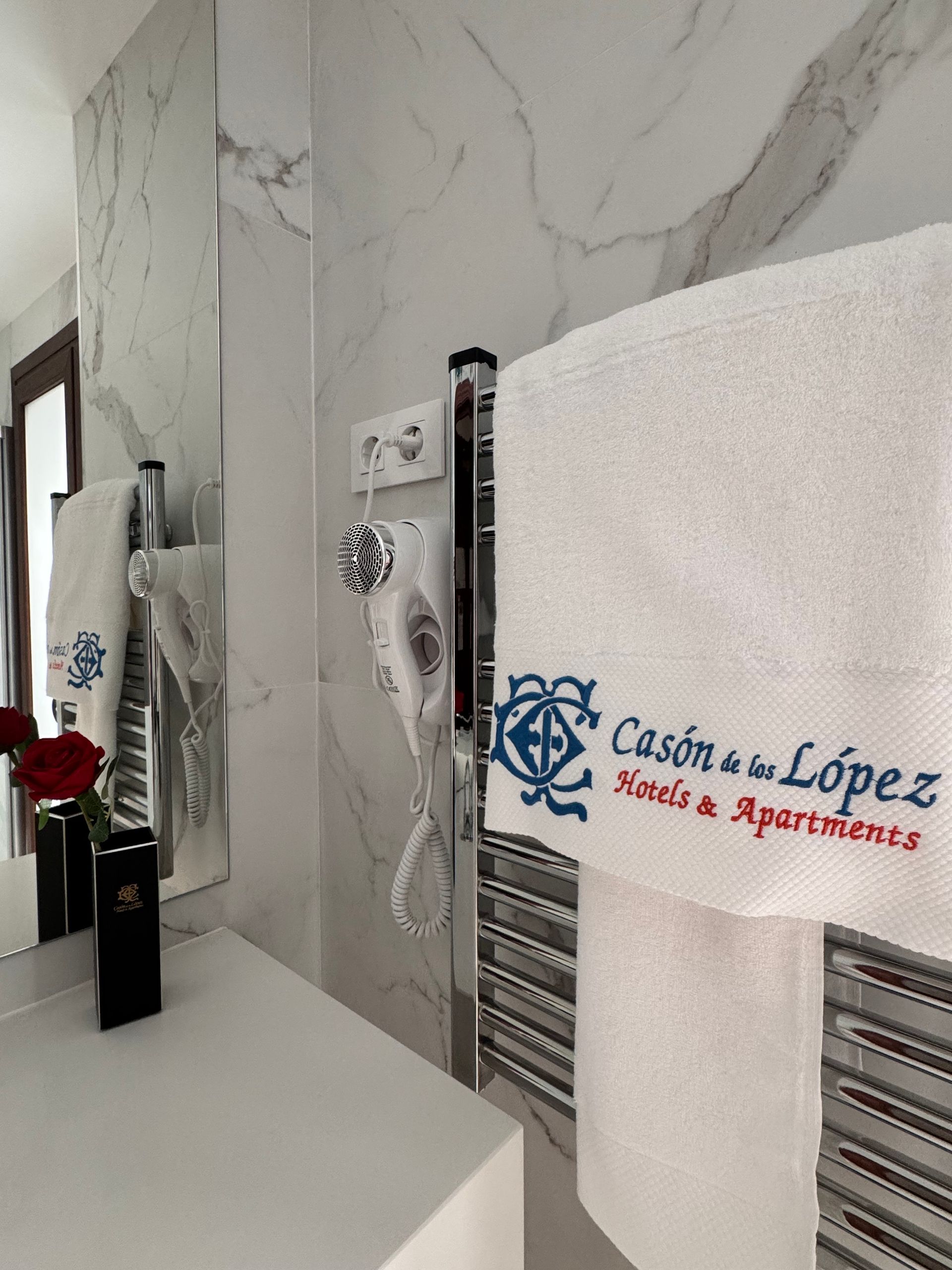 Un baño de hotel con una toalla blanca con el logotipo de Cason de los López en azul y rojo, un secador de pelo y un jarrón con una rosa.