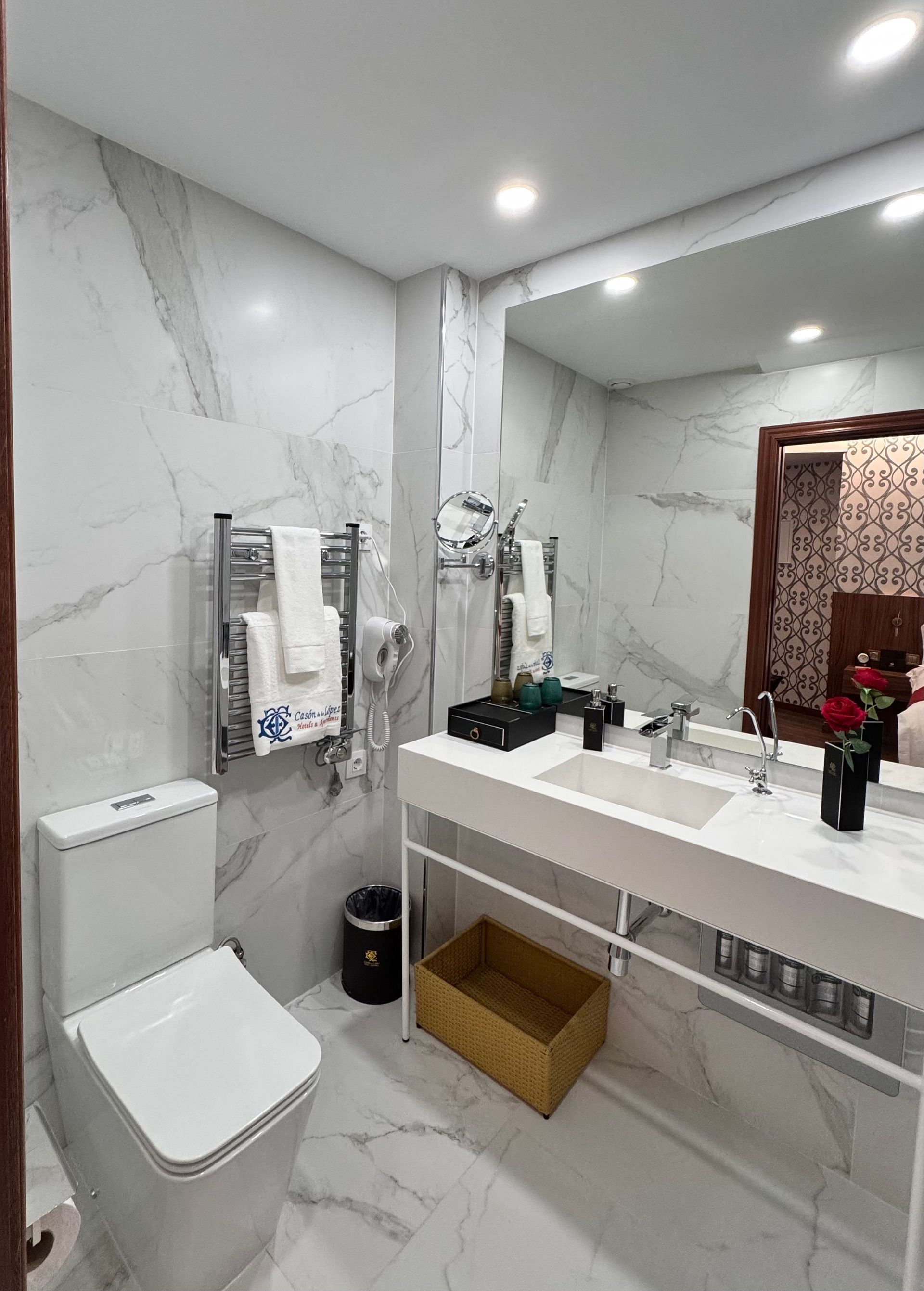 Un moderno baño con azulejos de mármol blanco, que cuenta con un elegante lavabo mural, un inodoro y un toallero eléctrico con toallas blancas.