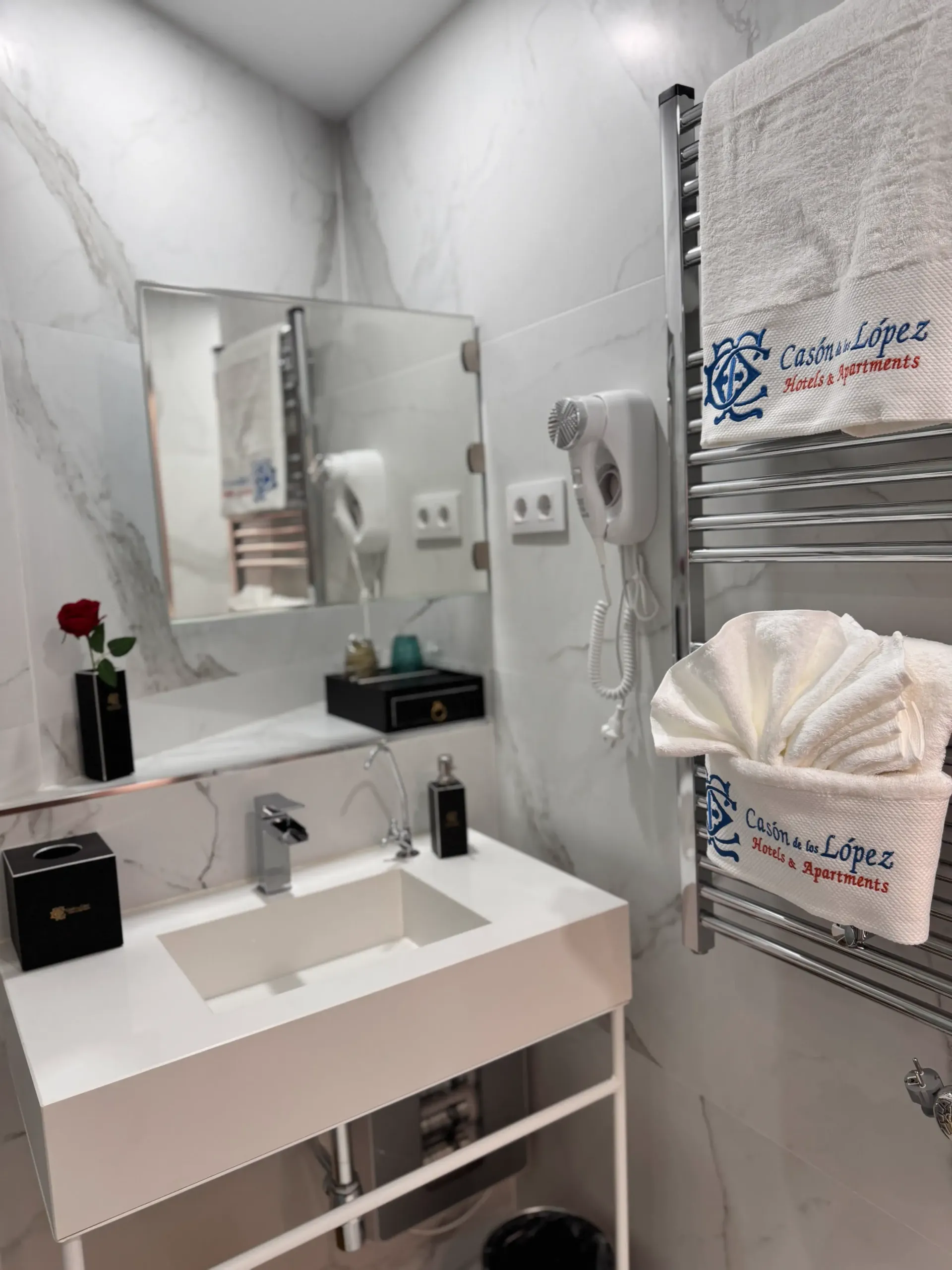 Un moderno lavabo blanco con paredes de mármol, un secador de pelo montado en la pared y un toallero eléctrico con toallas blancas dobladas.