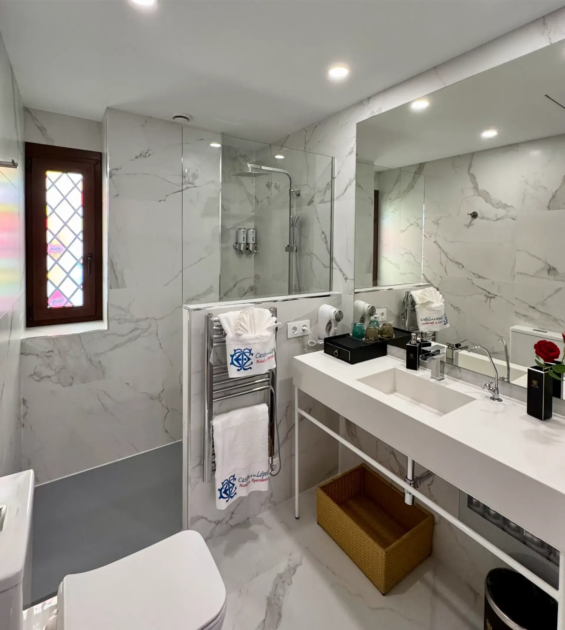 Un baño moderno con azulejos que imitan el mármol blanco, una ducha a ras de suelo con mampara de cristal, un lavabo con tocador y un mueble de almacenamiento de madera.