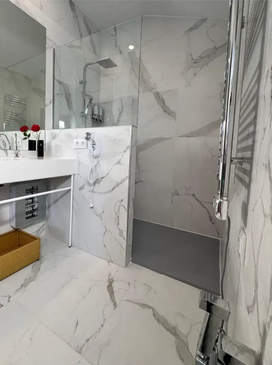 Un baño moderno con azulejos de mármol blanco, una ducha con mampara de cristal, un lavabo con marco de metal y un toallero eléctrico cromado.