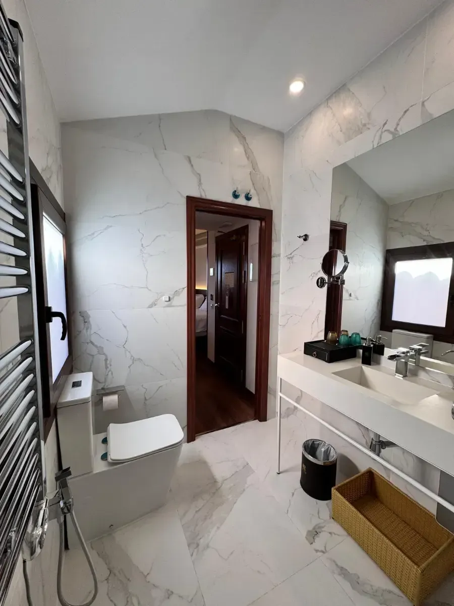 Un baño moderno con paredes de mármol blanco, inodoro, tocador suspendido con lavabo, espejo y puerta que da a un pasillo.