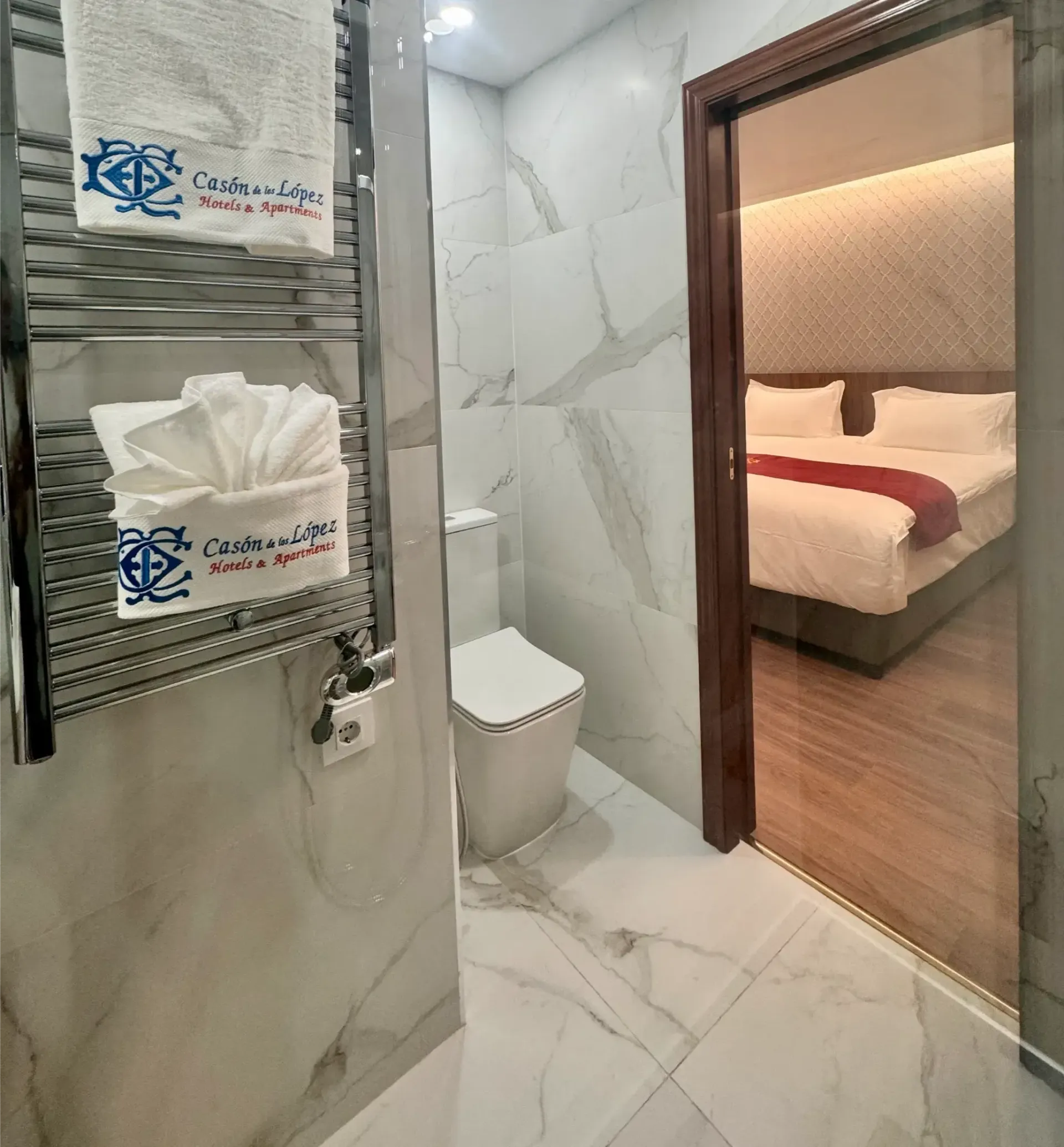 Un baño de hotel con un toallero con toallas dobladas, un inodoro y una puerta abierta que da a un dormitorio.