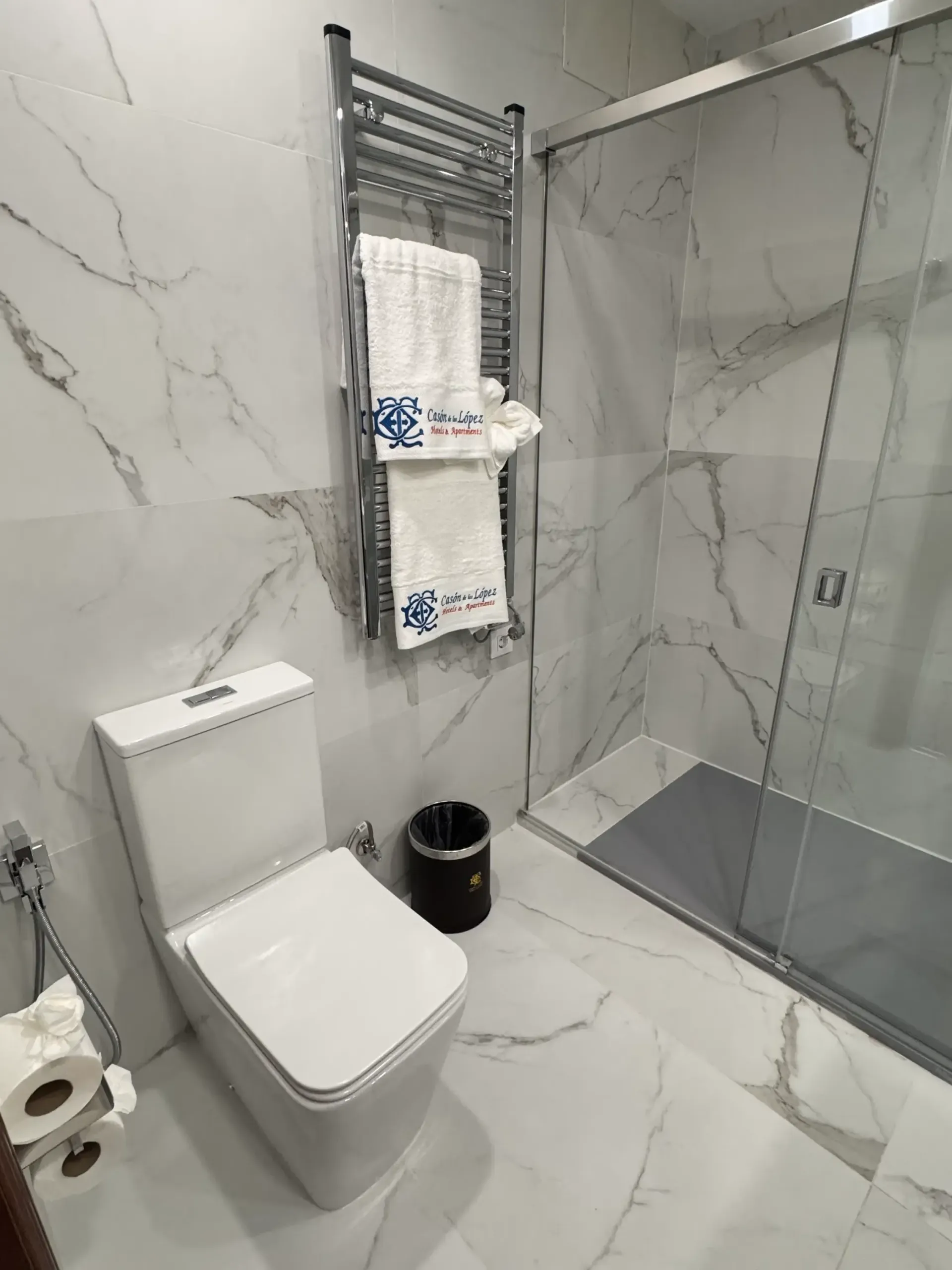 Baño moderno de mármol blanco con inodoro, toallero calefactable cromado con toallas colgantes, papelera negra y ducha de cristal.