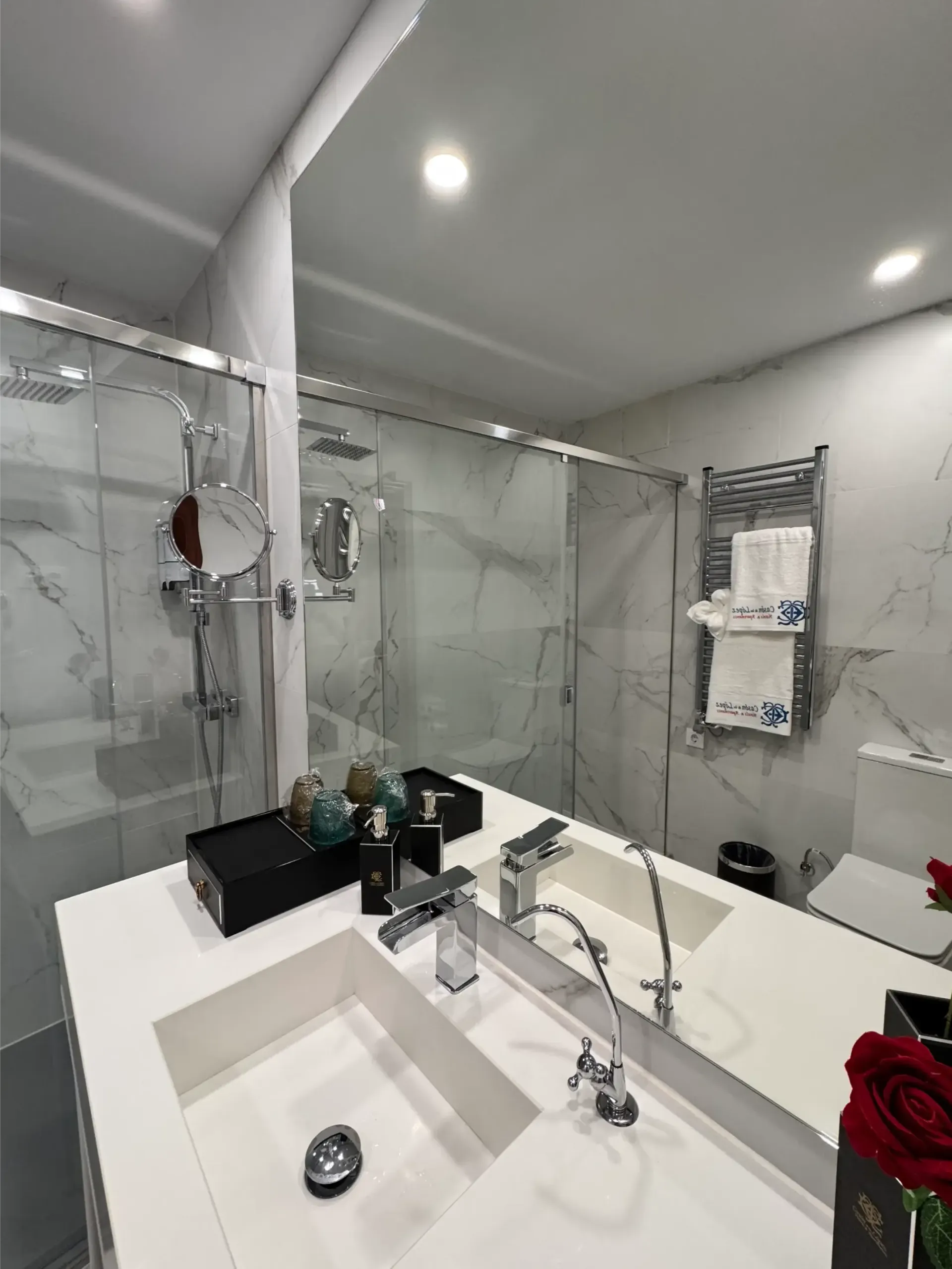 Un baño moderno con lavabo doble, paredes revestidas de mármol, cabina de ducha de cristal y toallero eléctrico de pared.