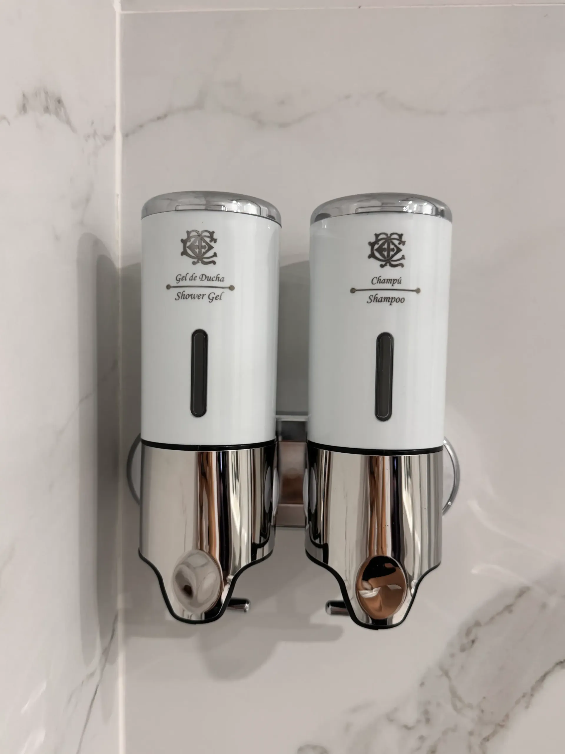 Dos dispensadores de jabón blancos y cromados, montados en la pared y con texto etiquetado, fijados a una pared revestida de mármol blanco.