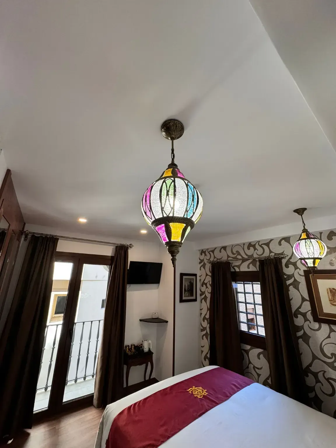 Dos coloridas lámparas colgantes de cristal iluminan una habitación de hotel con papel pintado estampado y cortinas oscuras.