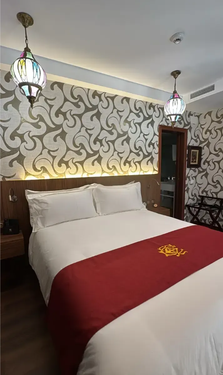 Una habitación de hotel con una cama tamaño queen, sábanas blancas y una alfombra roja, papel tapiz ornamentado y dos lámparas colgantes de cristal.