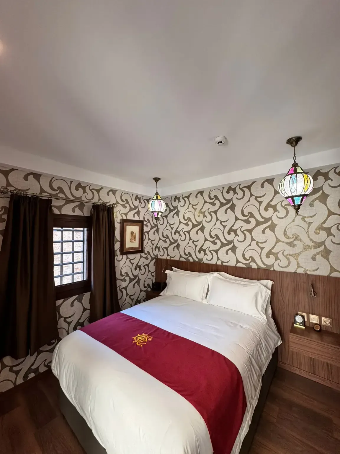 Un dormitorio acogedor con papel pintado estampado, una cama con una estera roja, cortinas marrones y dos lámparas colgantes decorativas.