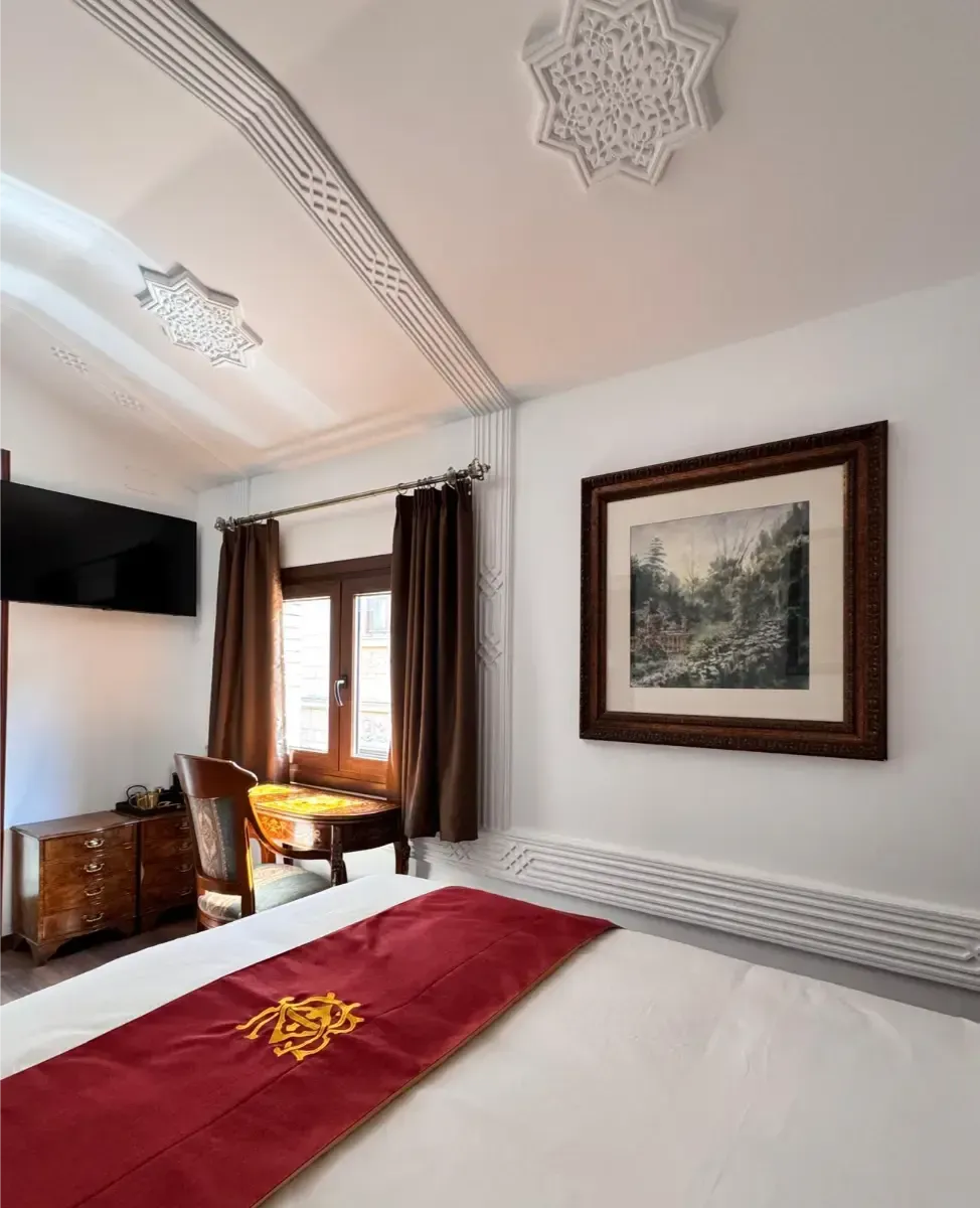 Un dormitorio con una cama con una alfombra roja, un escritorio de madera con una silla, un televisor montado en la pared y un cuadro de paisaje enmarcado.