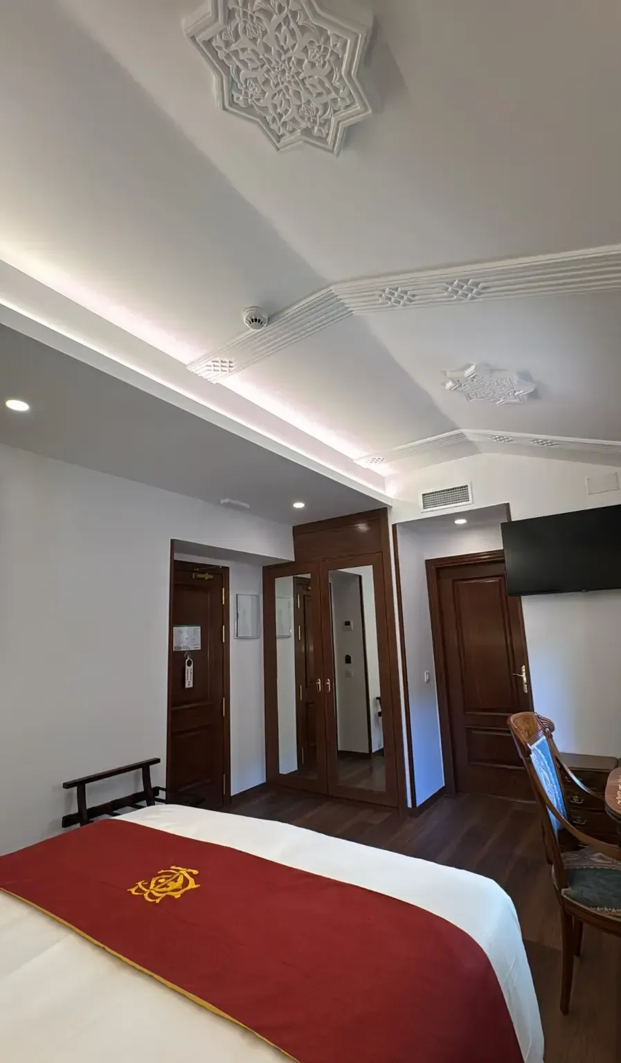 Una habitación de hotel moderna con una cama con una senda roja, puertas de madera, un televisor montado en la pared y un intrincado detalle en el techo.