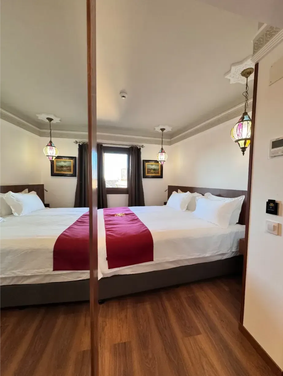 Una habitación de hotel con dos camas individuales con sábanas blancas y caminos de mesa rojos, suelo de madera y faroles colgantes decorativos.