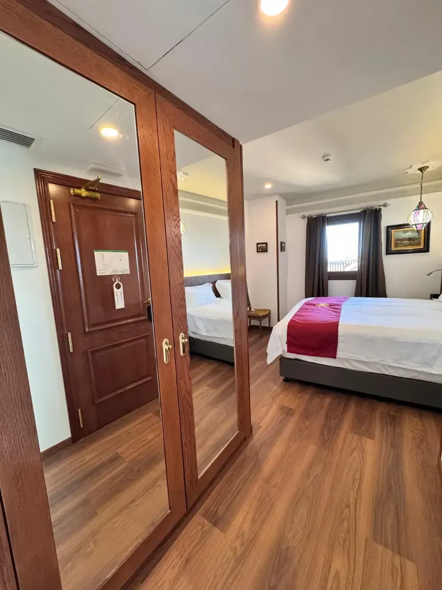 Una habitación de hotel con un gran espejo en primer plano que refleja una cama, y ​​con vista a una segunda cama y una ventana al fondo.