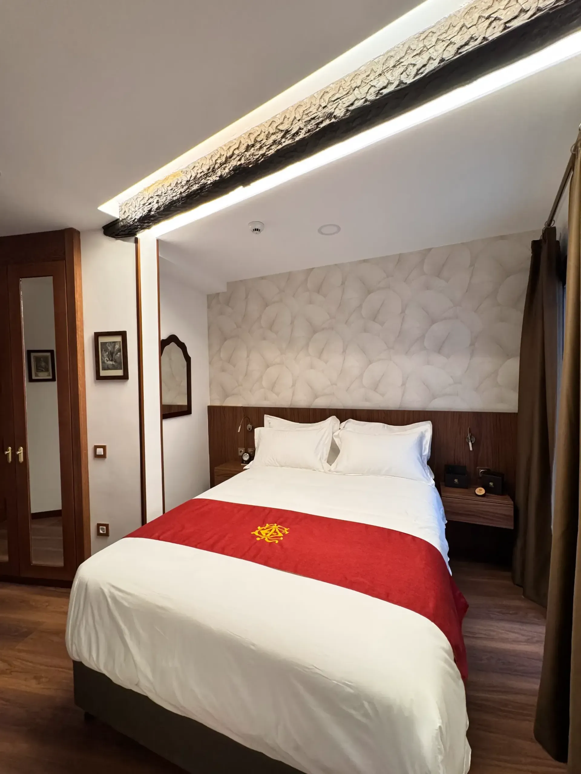 Una habitación de hotel con una cama doble vestida con sábanas blancas, una alfombra roja y papel pintado con un estampado claro sobre el cabecero.