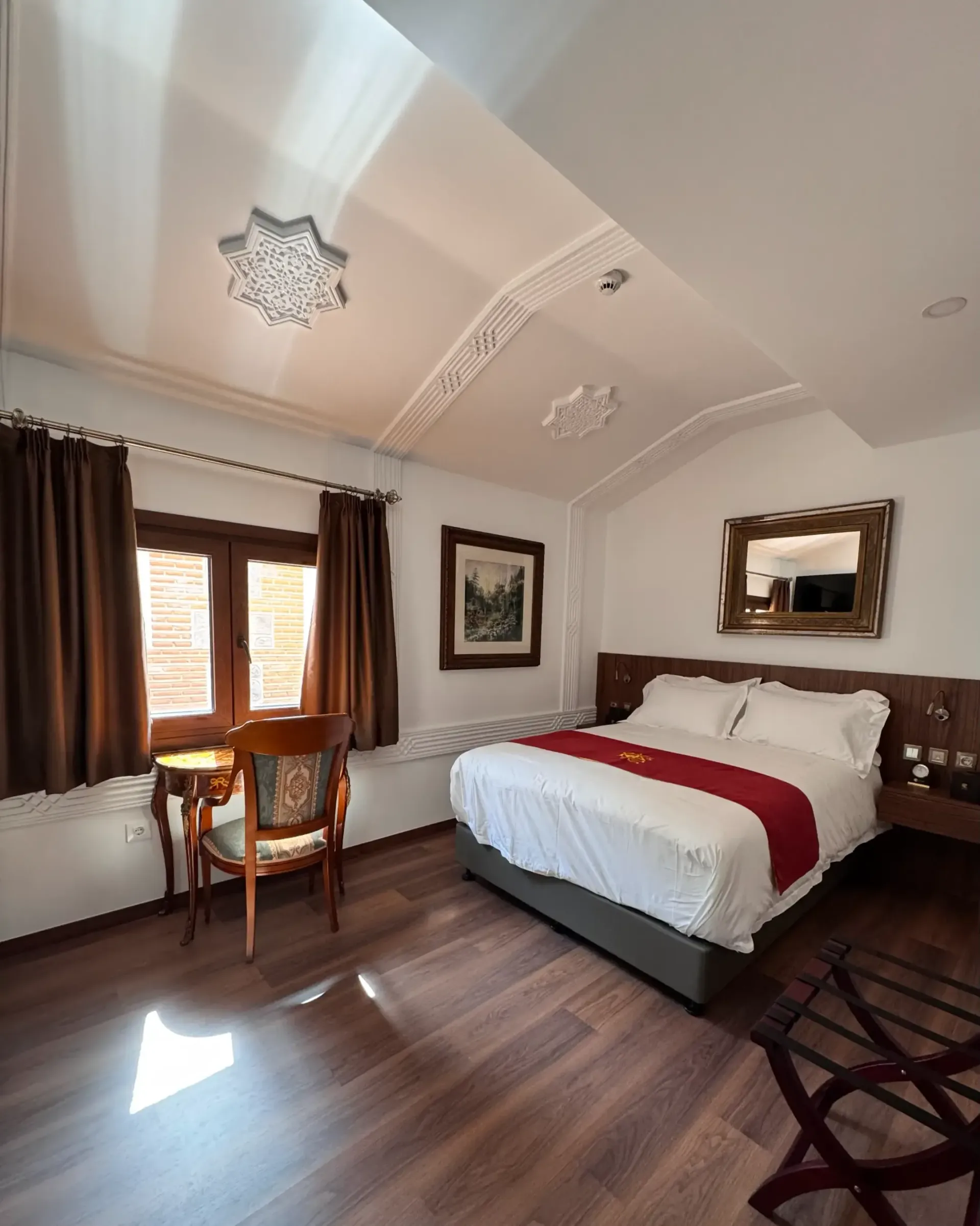 Una habitación de hotel con una cama de matrimonio, muebles de madera oscura, cortinas marrones y detalles ornamentales en el techo.