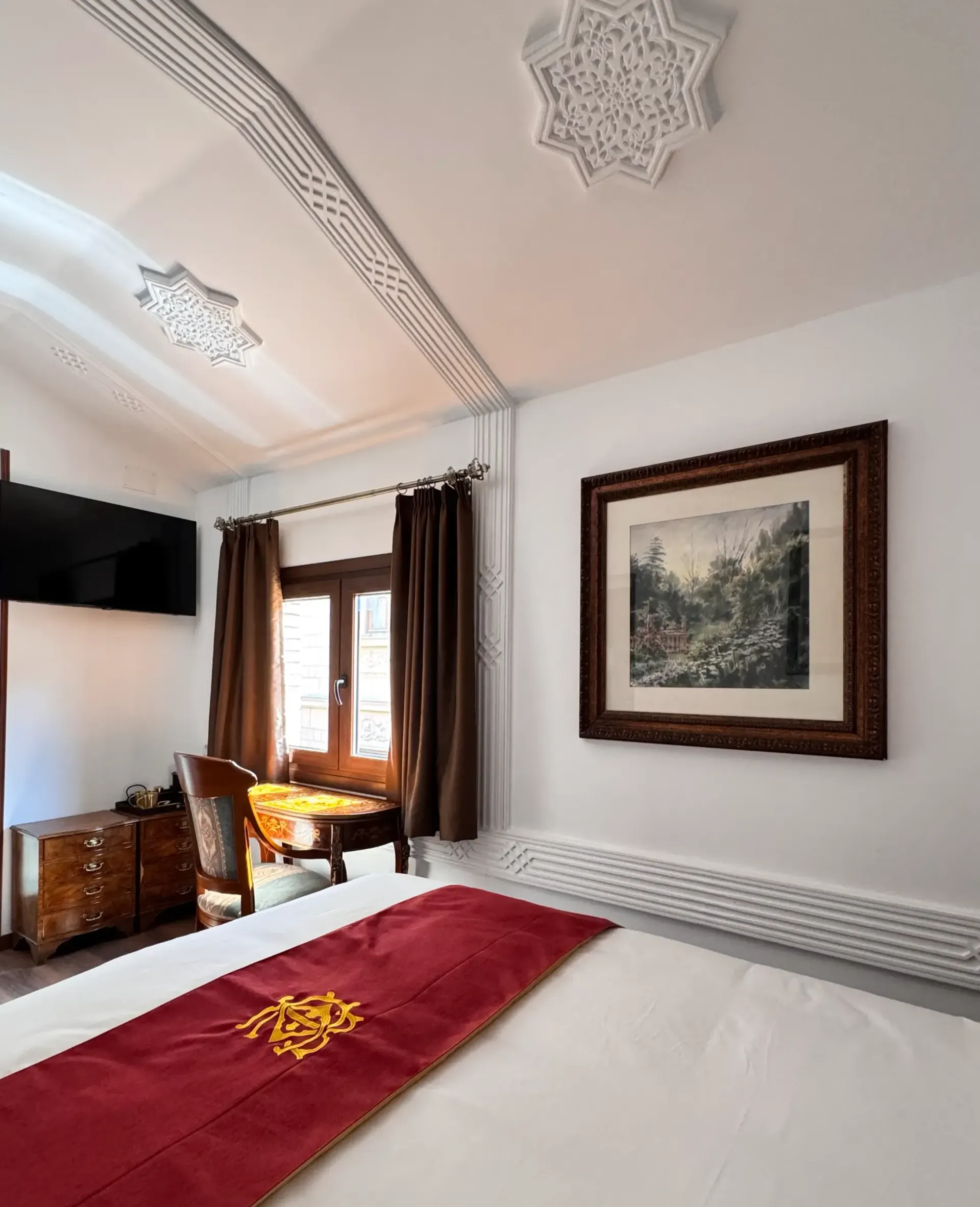 Un dormitorio con una cama con una alfombra roja, un escritorio junto a una ventana con cortinas marrones y un cuadro enmarcado en la pared.