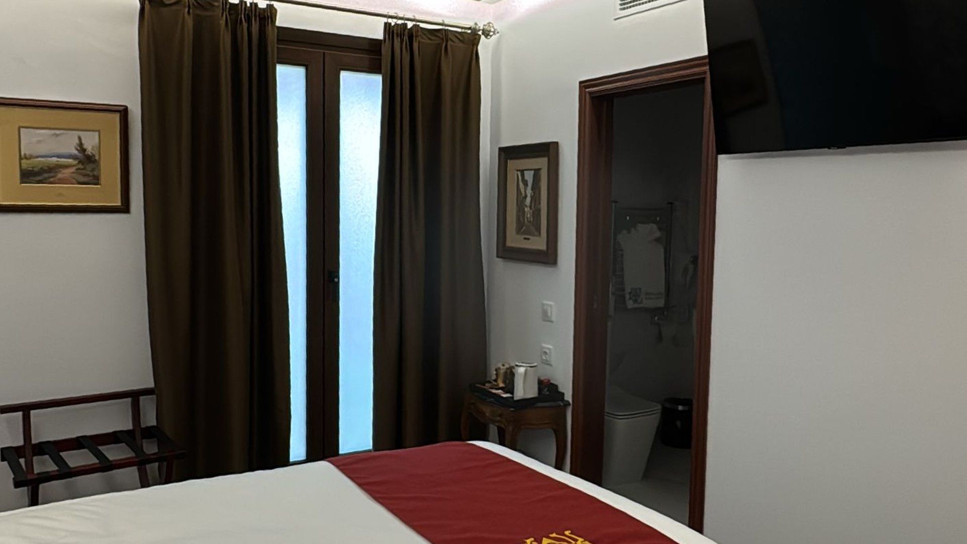 Una habitación de hotel con una cama con una estera roja, una ventana con cortinas marrones, obras de arte en las paredes y un baño abierto.