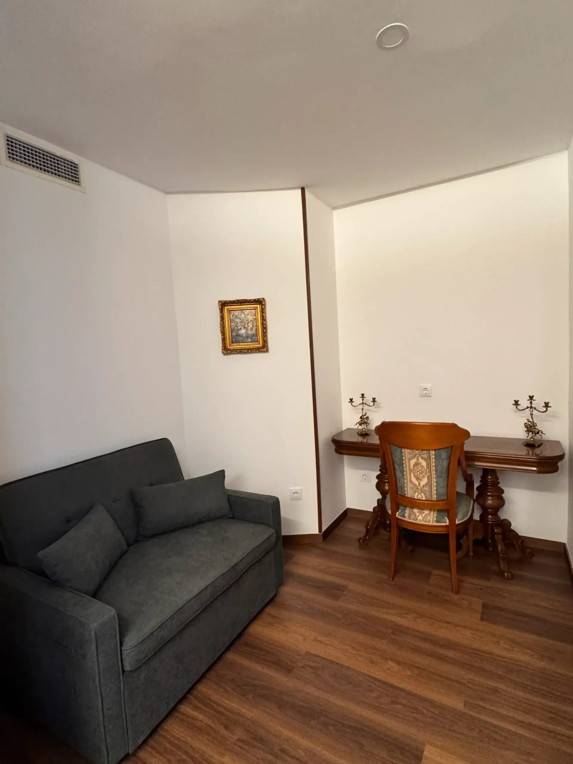 Una pequeña habitación con un sofá gris, un escritorio de madera con una silla a juego y un cuadro enmarcado en la pared blanca.