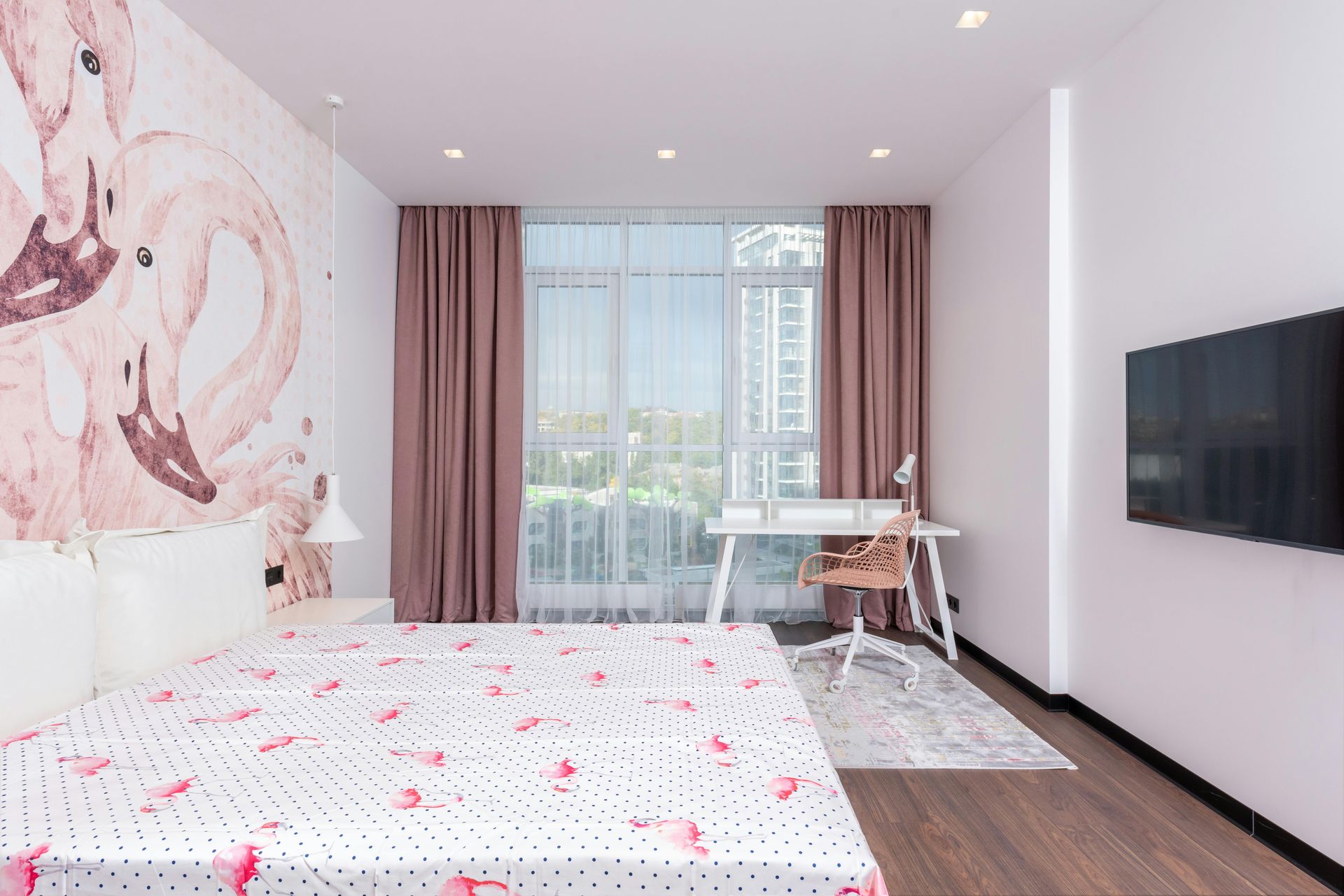 A bedroom with a bed , desk , television and flamingos on the wall.