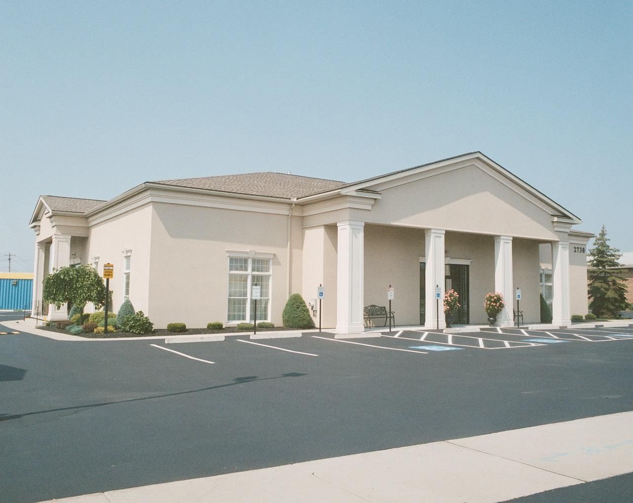Cream-colored building with white columns and brown roof, likely a funeral home. Asphalt parking lot in front.