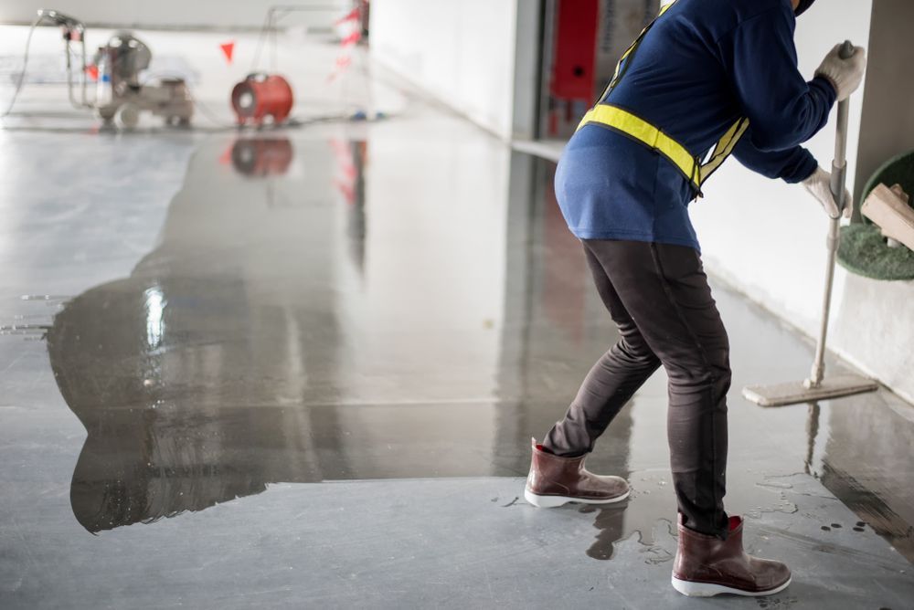Person Applying A Sealant To A Gray Floor In A Warehouse Setting — TMP Painting Services in Beaconsfield, QLD