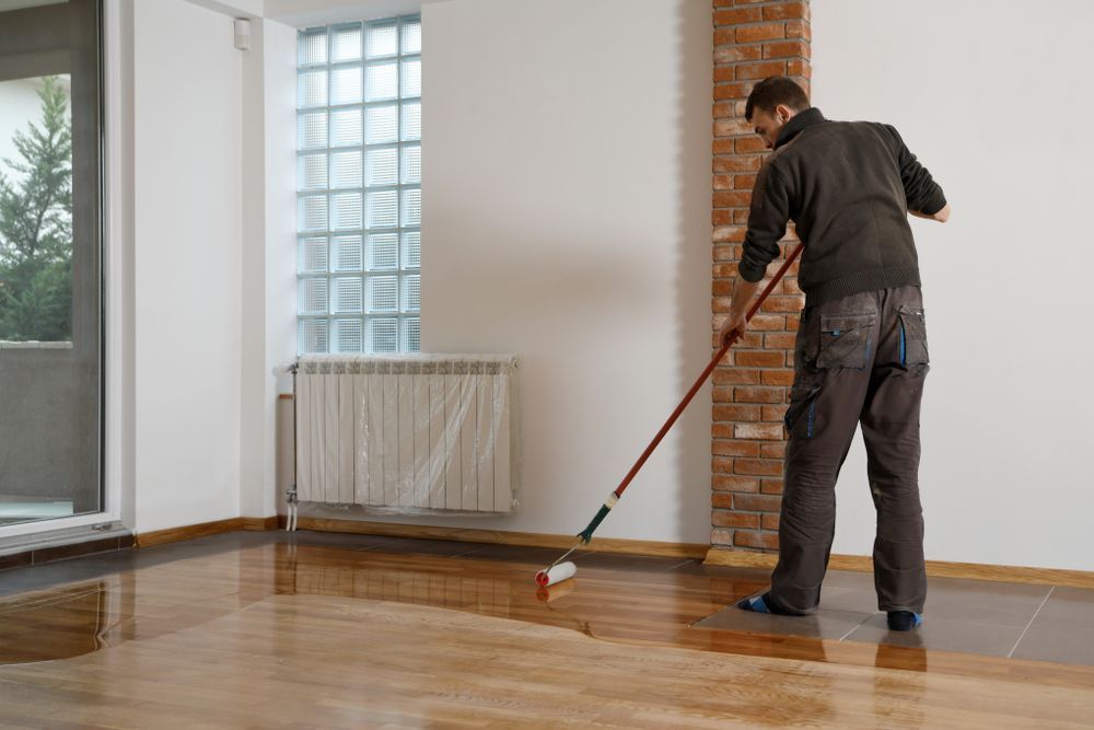 Man Applying Varnish To A Wooden Floor In A Room With White Walls — TMP Painting Services in Beaconsfield, QLD