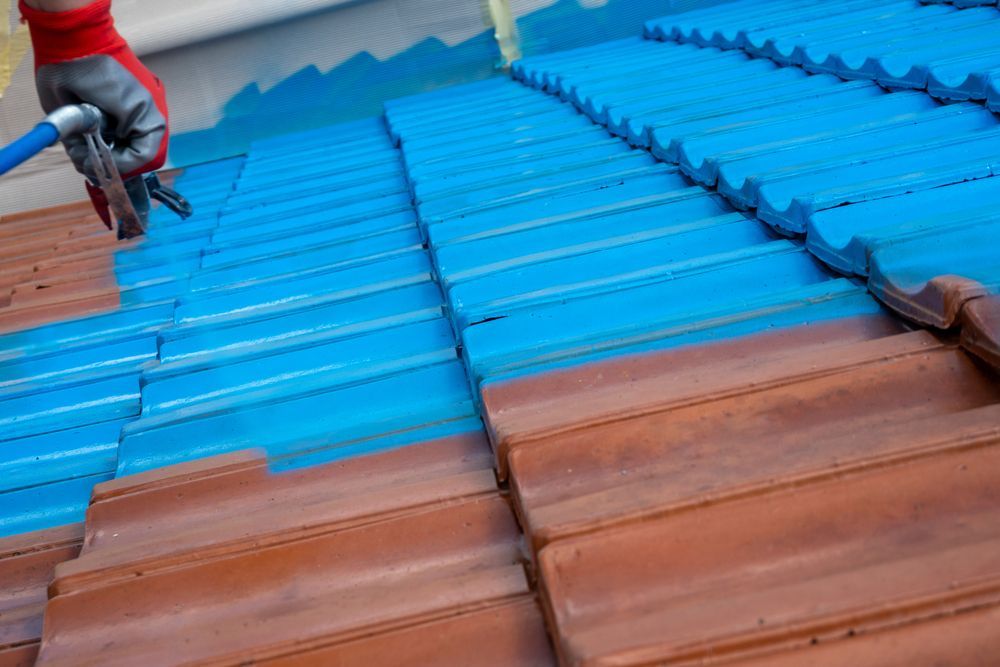 Blue paint being sprayed on a tiled roof by a person wearing gloves — TMP Painting Services in Paget, QLD