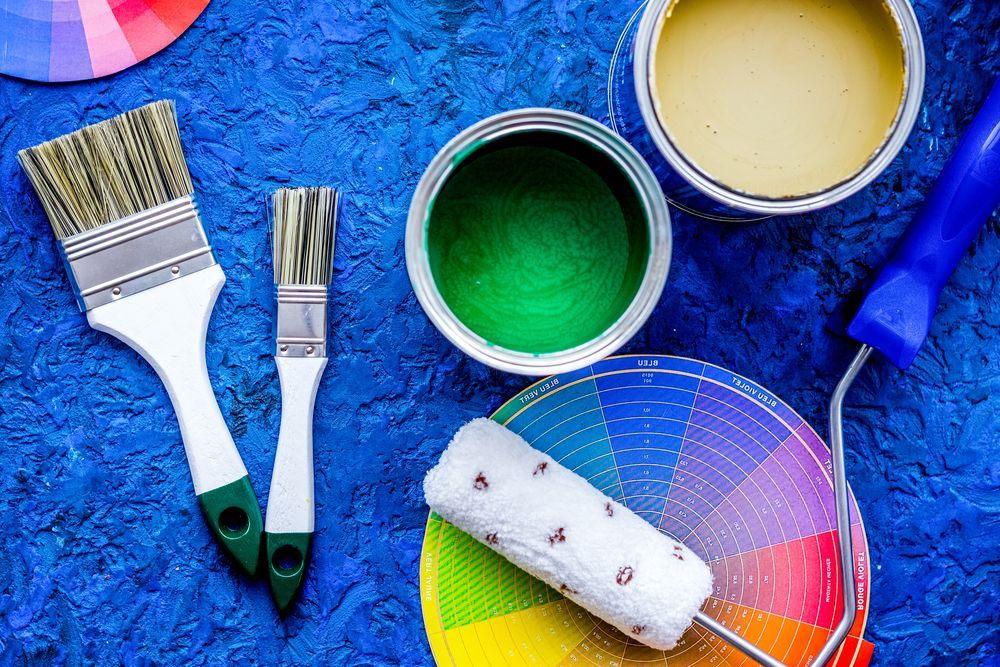 Paint supplies on blue surface: brushes, paint cans (green, yellow), color swatches, paint roller — TMP Painting Services in Sarina, QLD