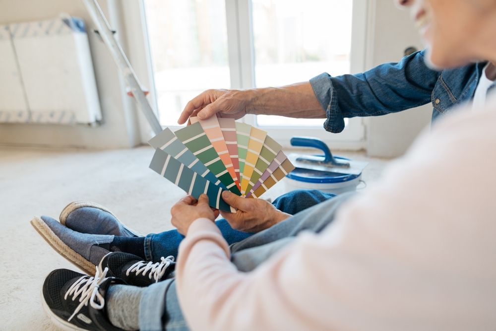 Couple holding paint swatches, deciding on wall color in a room with a window, paint roller, and a rug — TMP Painting Services in Beaconsfield, QLD