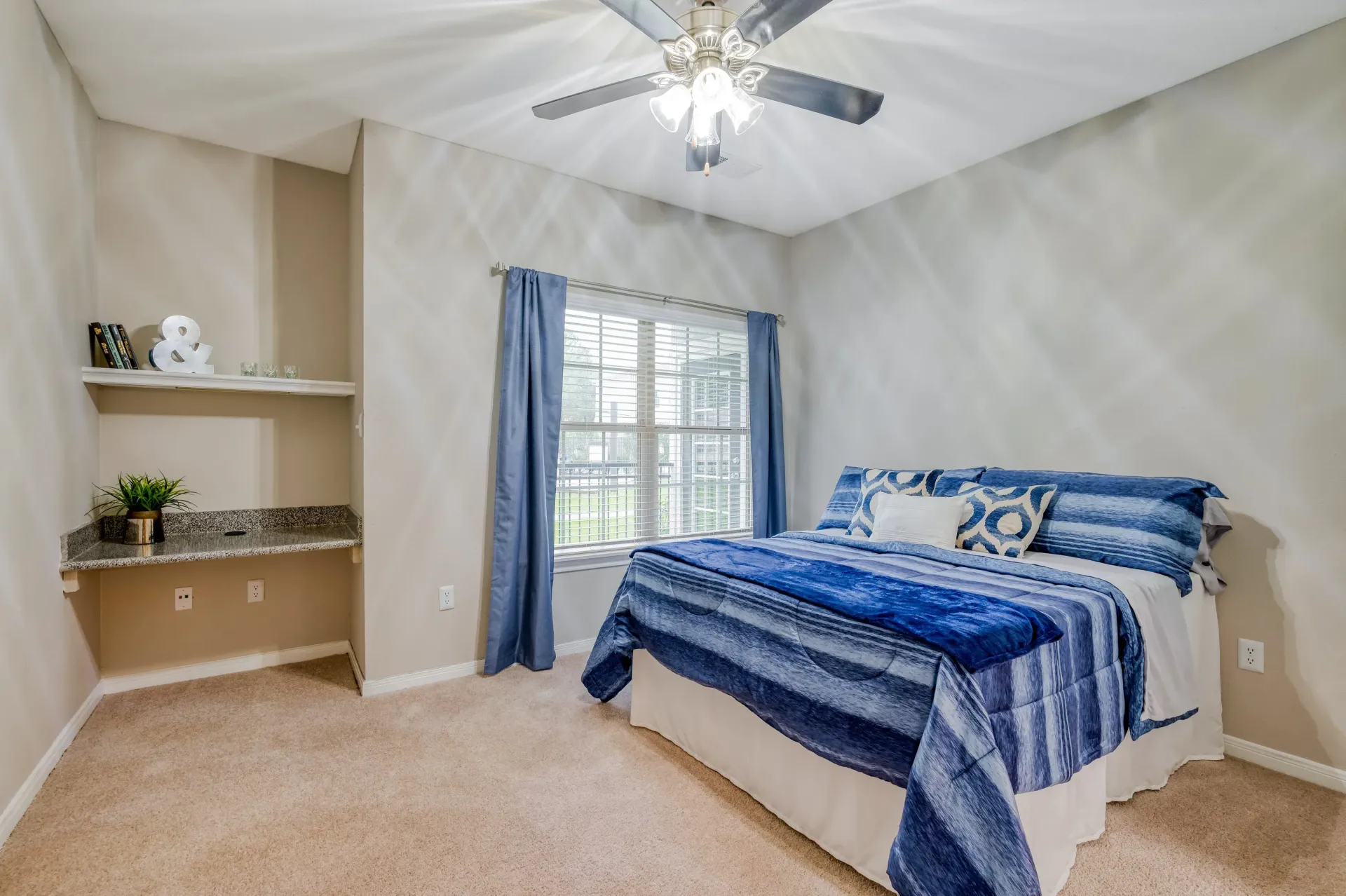 Bedroom with blue bedding, window with blue curtains, ceiling fan, and a built-in desk nook.